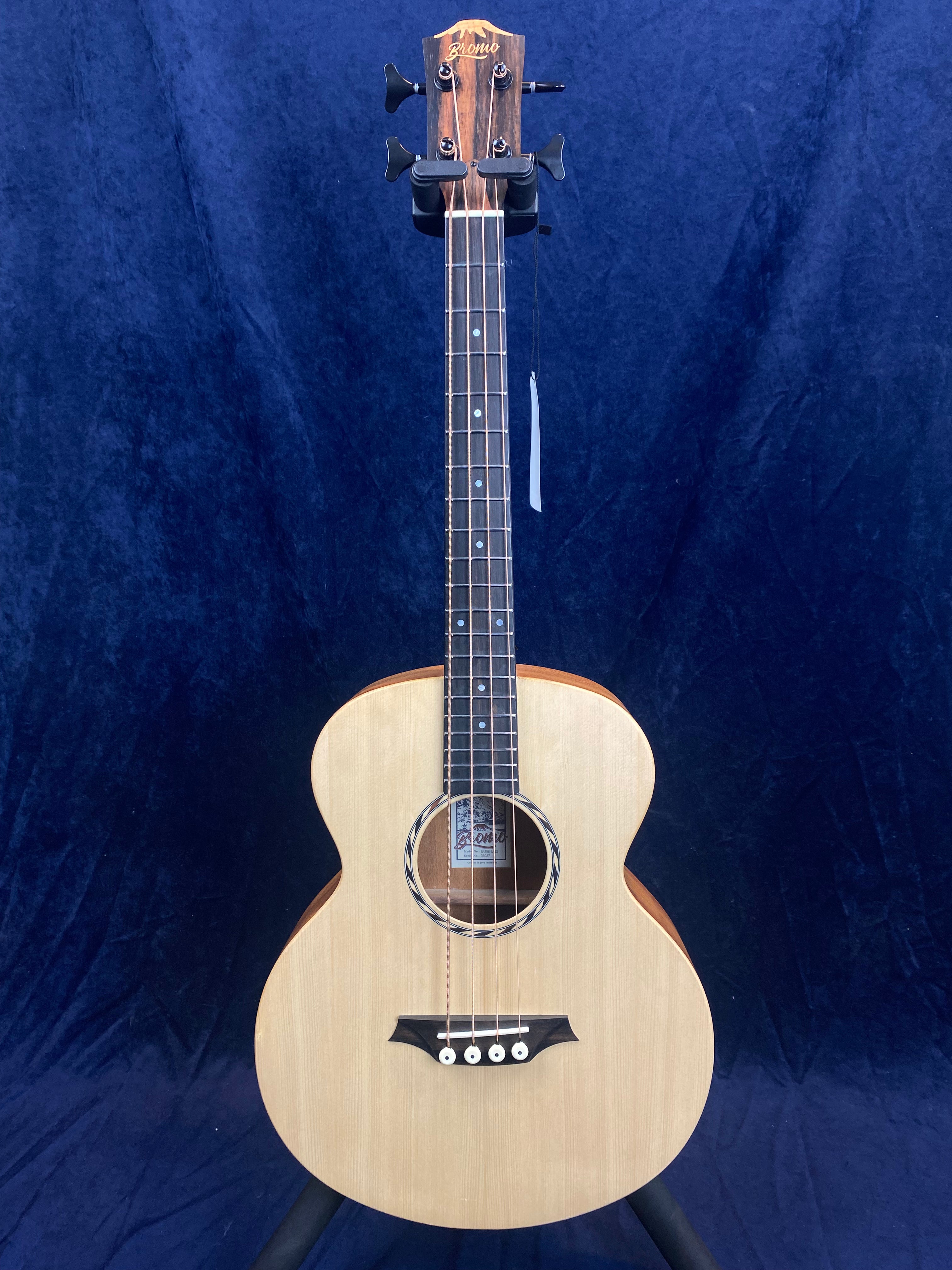 Acoustic guitar with a natural wood finish, rounded body, dark fretboard, and four tuning pegs on a wooden headstock against a dark blue background.