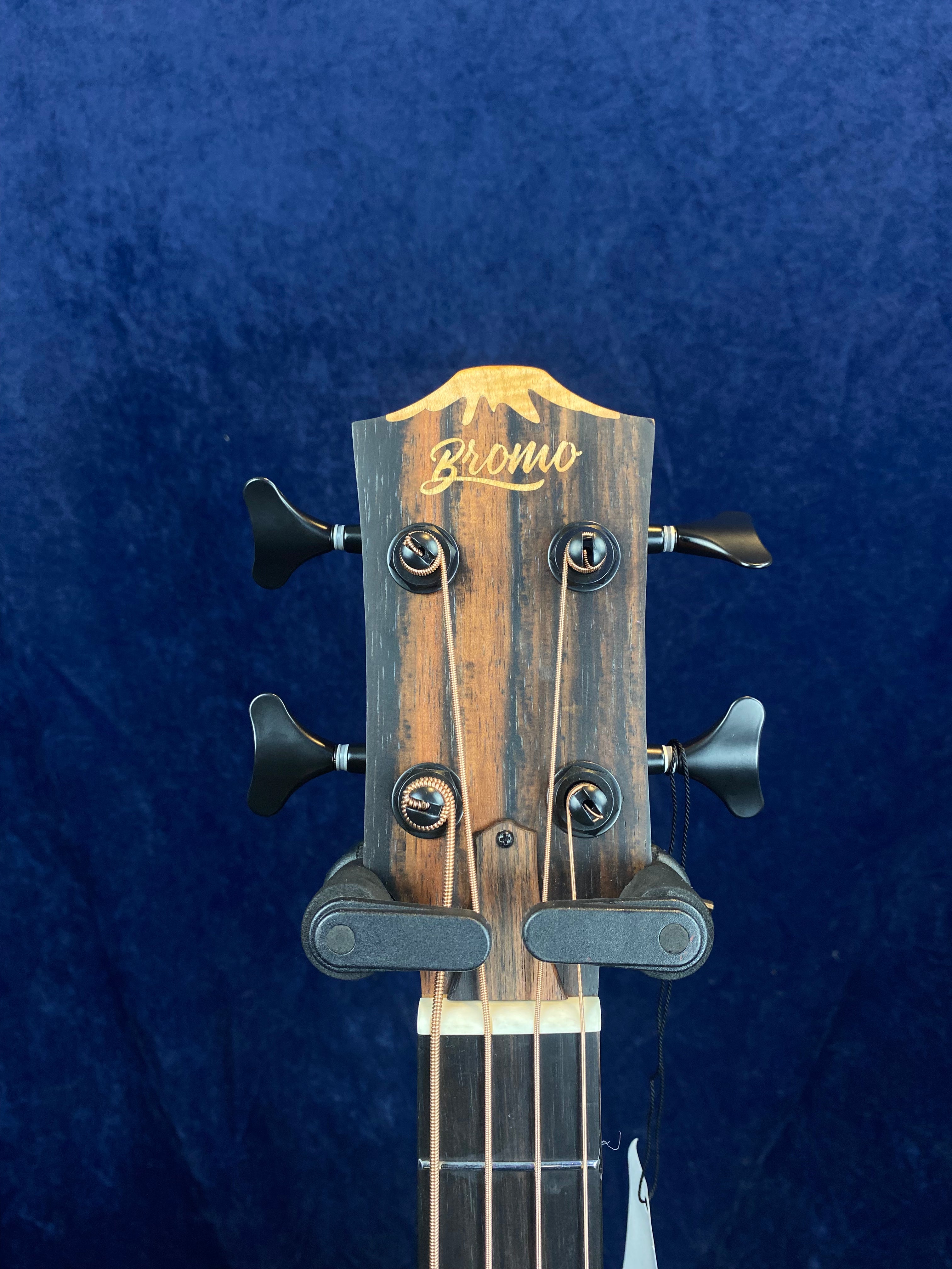 Close-up of an acoustic guitar headstock with the brand name "Bromo," four tuning pegs, and two string retainers, against a blue background.