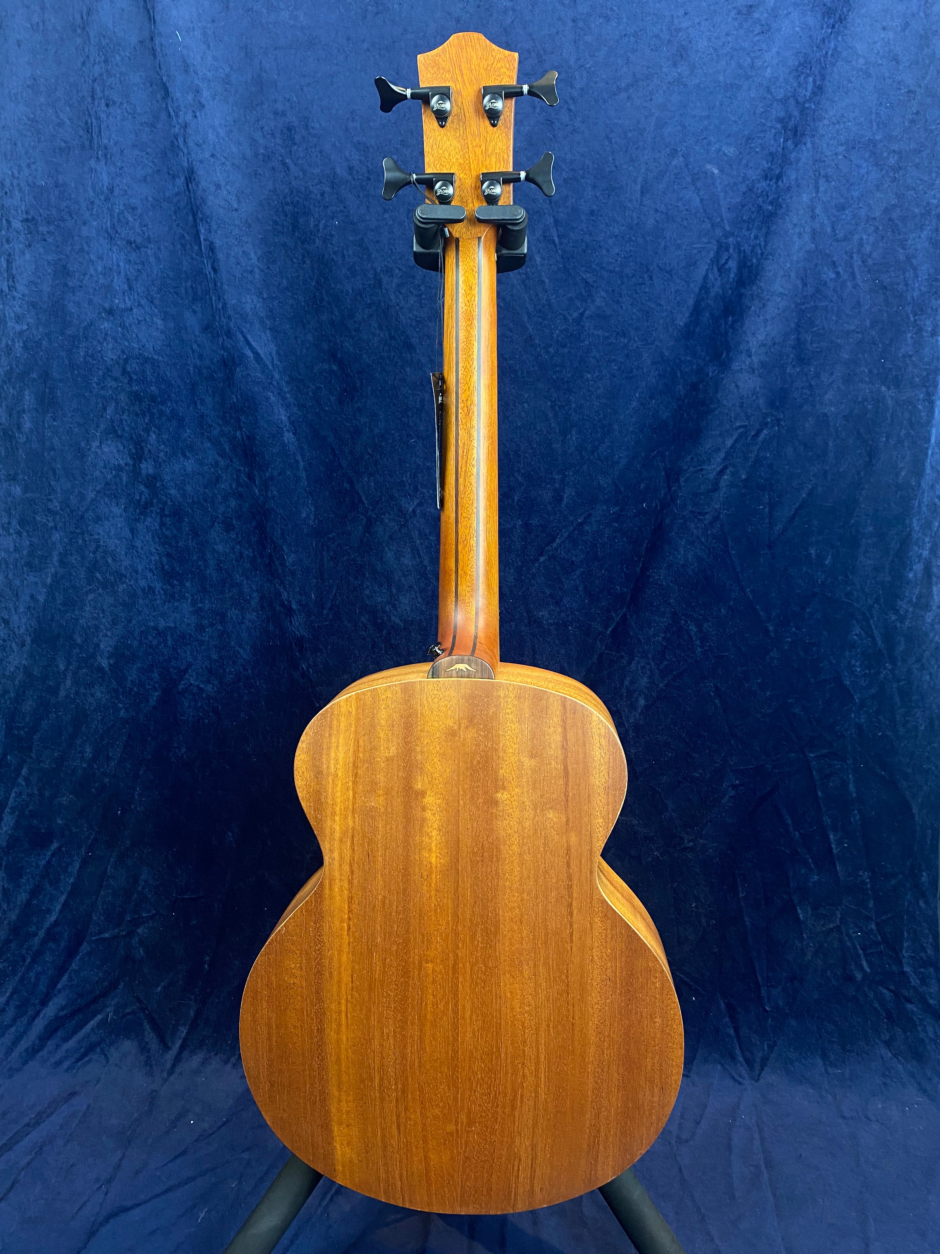 Acoustic guitar shown from the back, displaying smooth brown wood grain, four tuning pegs, and a curved body against a dark blue background.