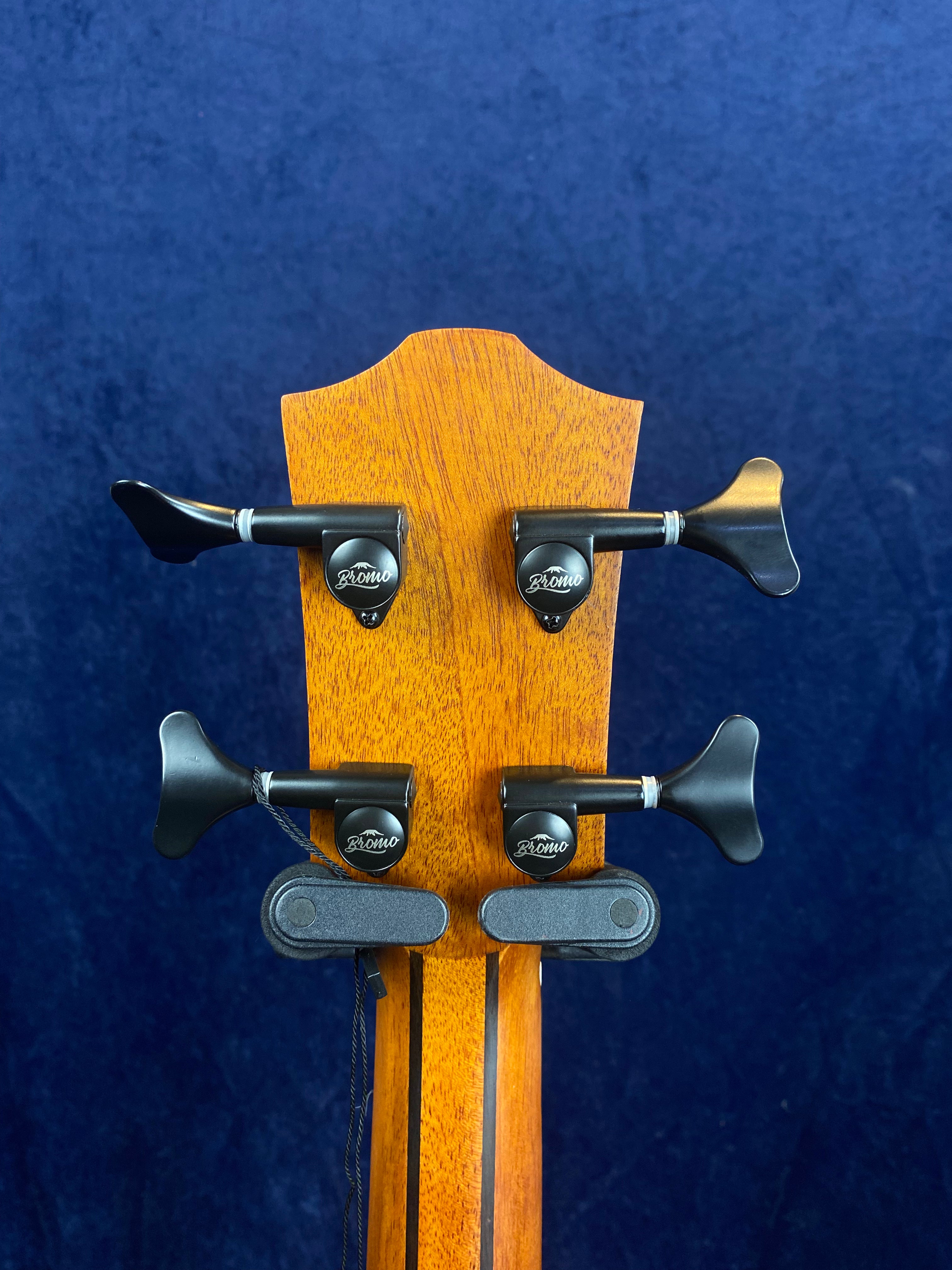 The back of a wooden acoustic guitar headstock with four black tuning pegs, each labeled “Bromo,” against a dark blue background.