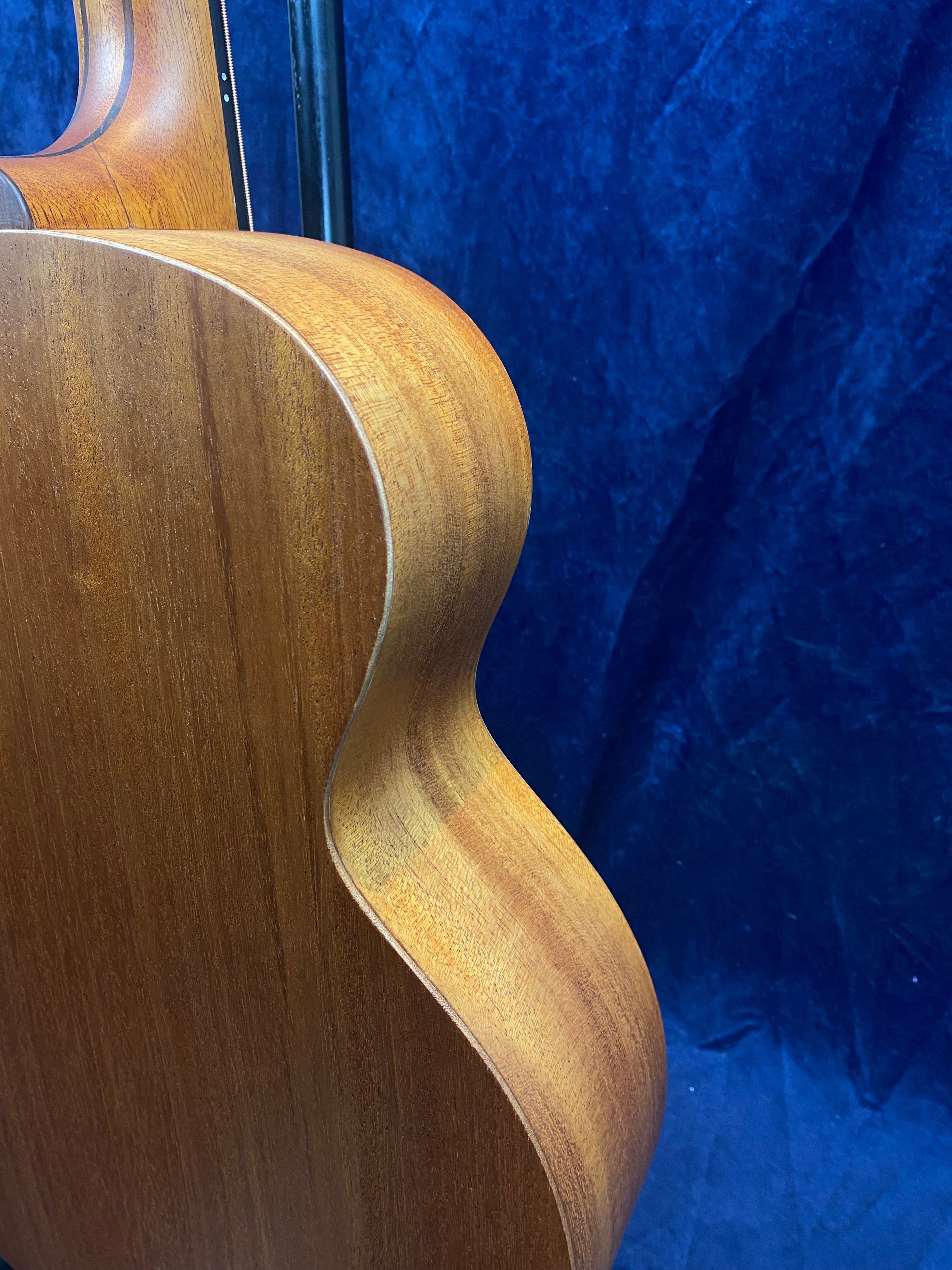 Side view of an acoustic guitar showing smooth, natural wood grain and gentle curves against a dark blue textured background.