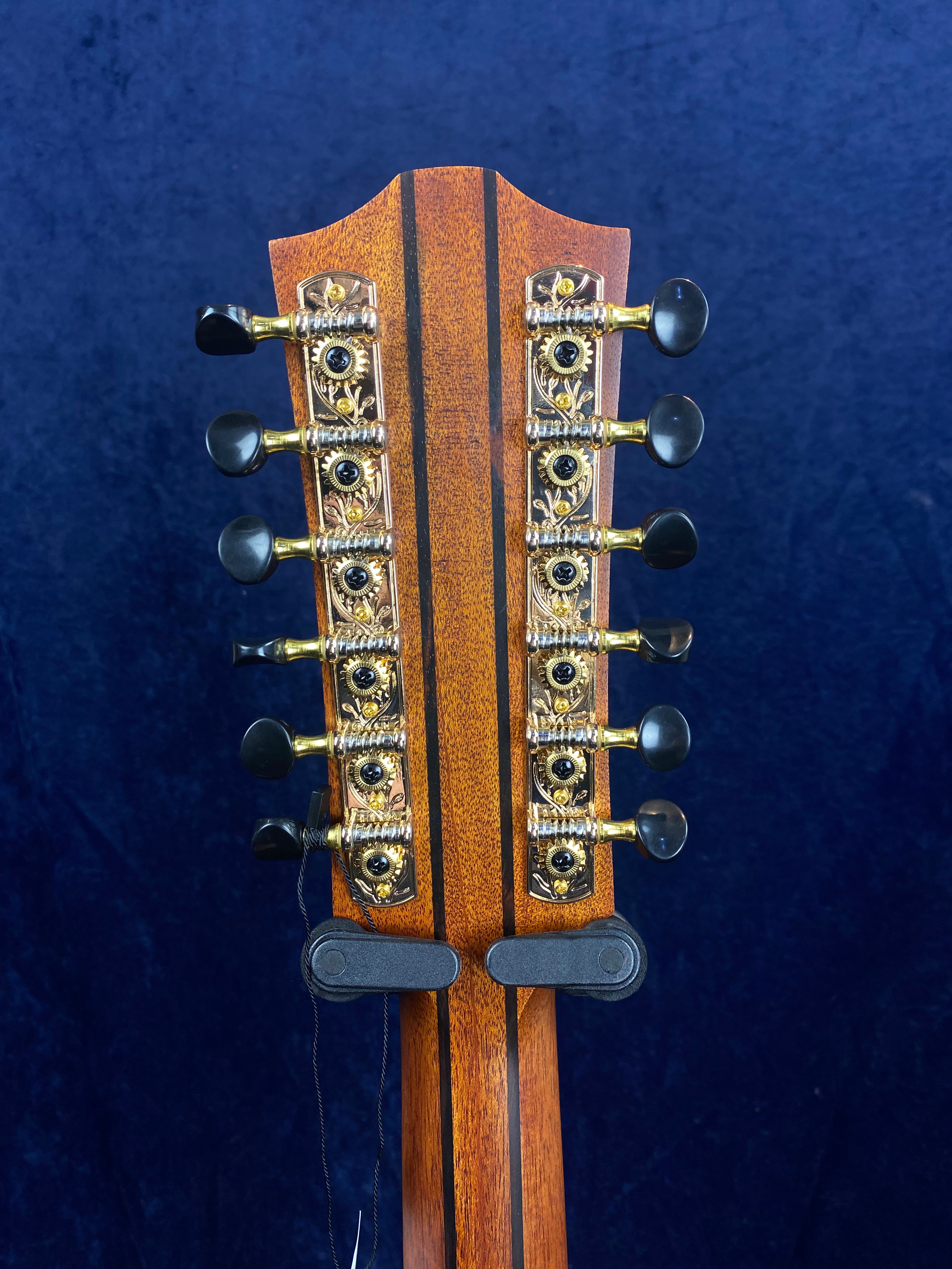 Back of a 12-string acoustic guitar headstock, showing ornate gold tuning machines with black knobs against a dark blue background.