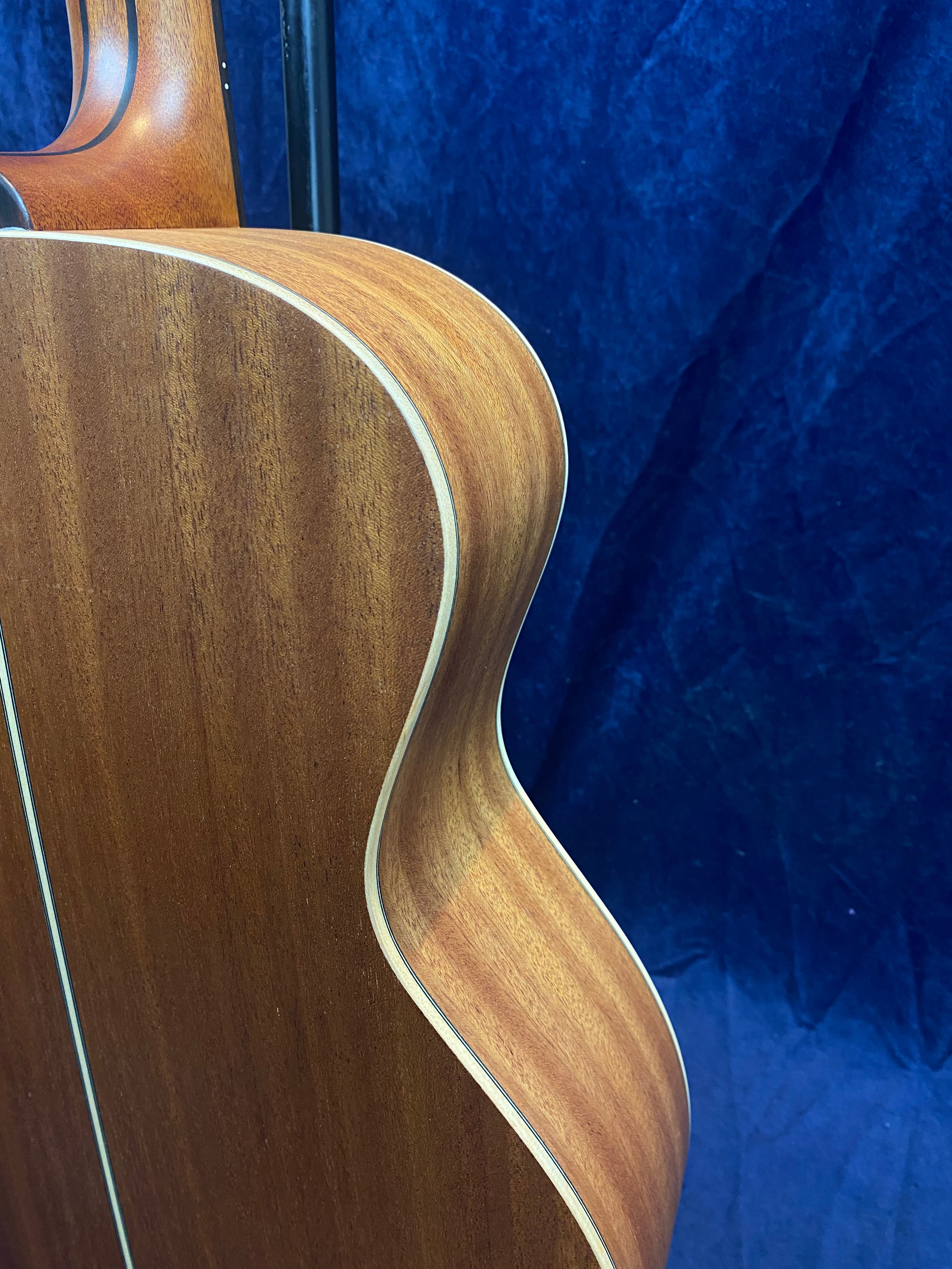 Curved side view of an acoustic guitar with natural wood finish and light binding, shown against a dark blue background.