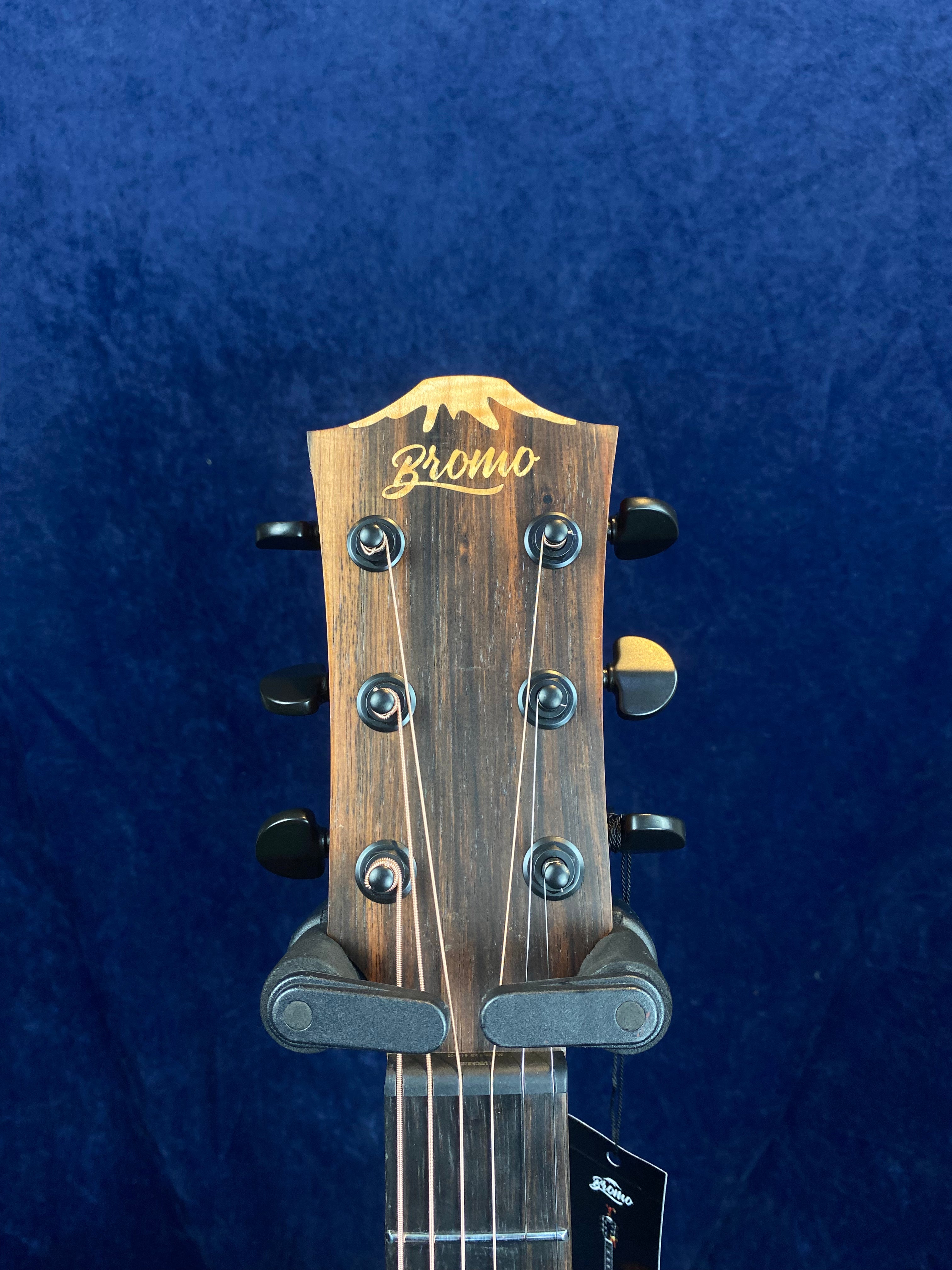 Dark wood acoustic guitar headstock with six tuning pegs and the brand name "Bromo" in gold script, set against a deep blue background.