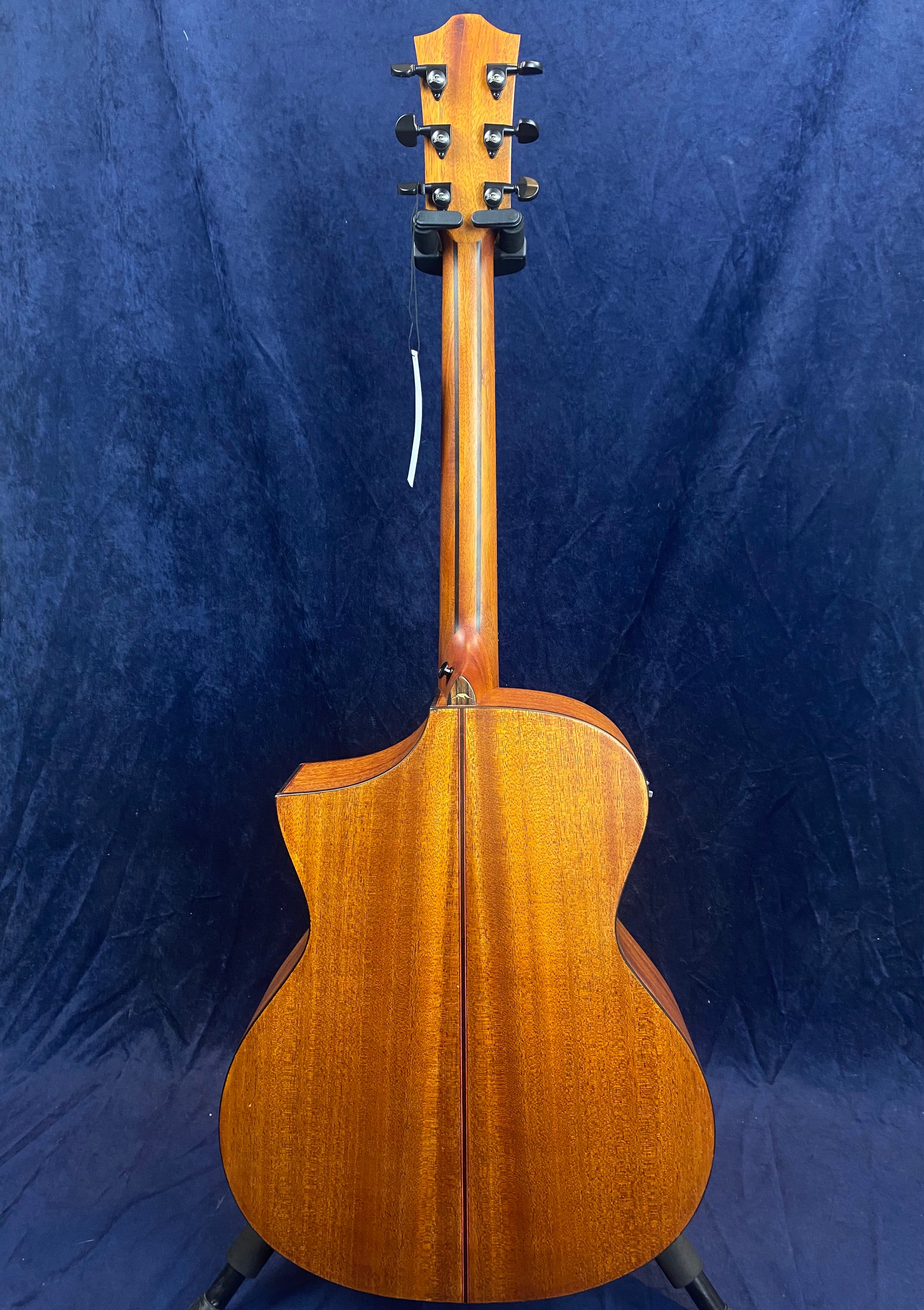 Acoustic guitar shown from the back, featuring a warm brown wood finish, cutaway body design, and six tuning pegs against a dark blue background.