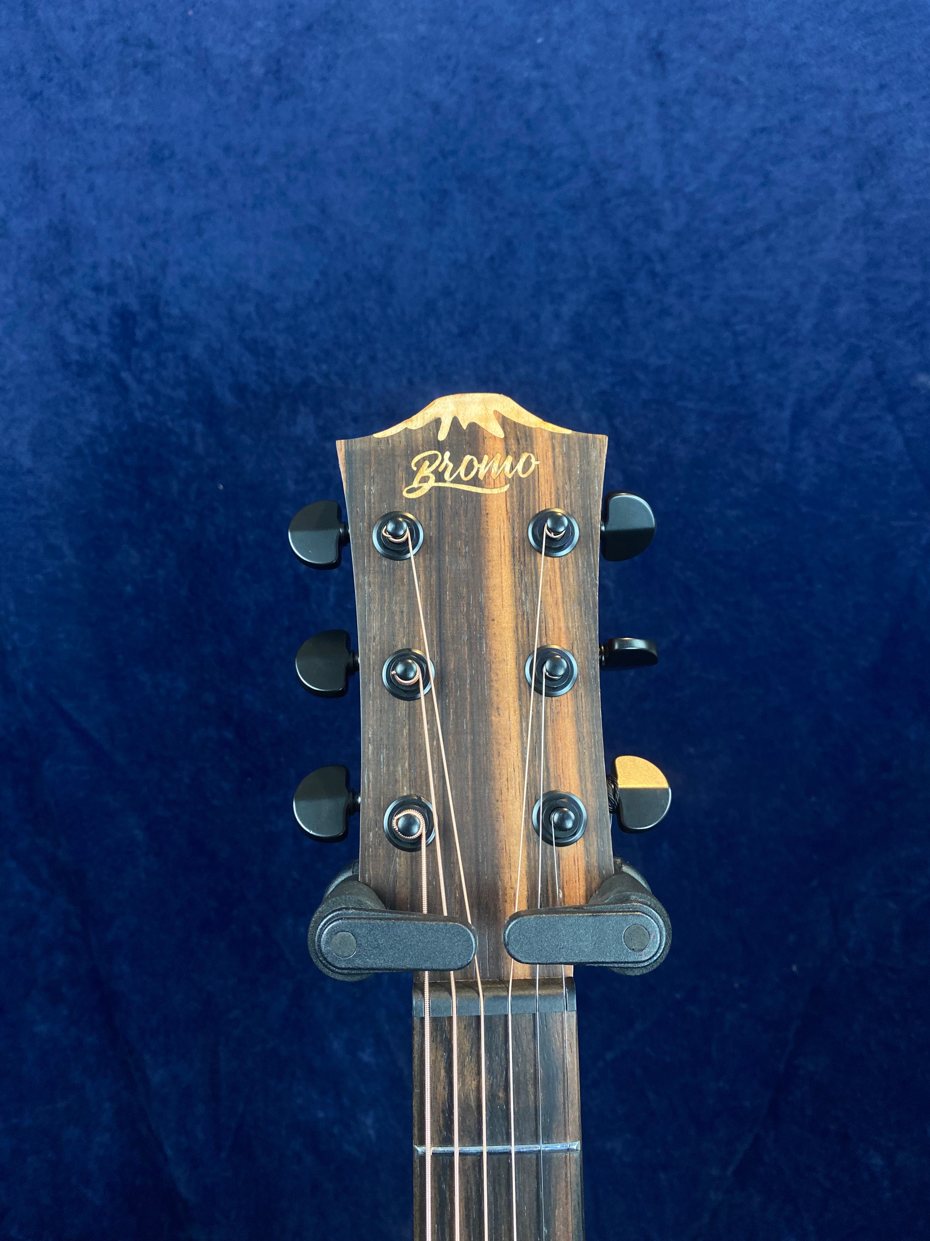 Tête de guitare acoustique en bois foncé avec six mécaniques noires, logo « Bromo » gravé sur le haut, fond bleu uni.