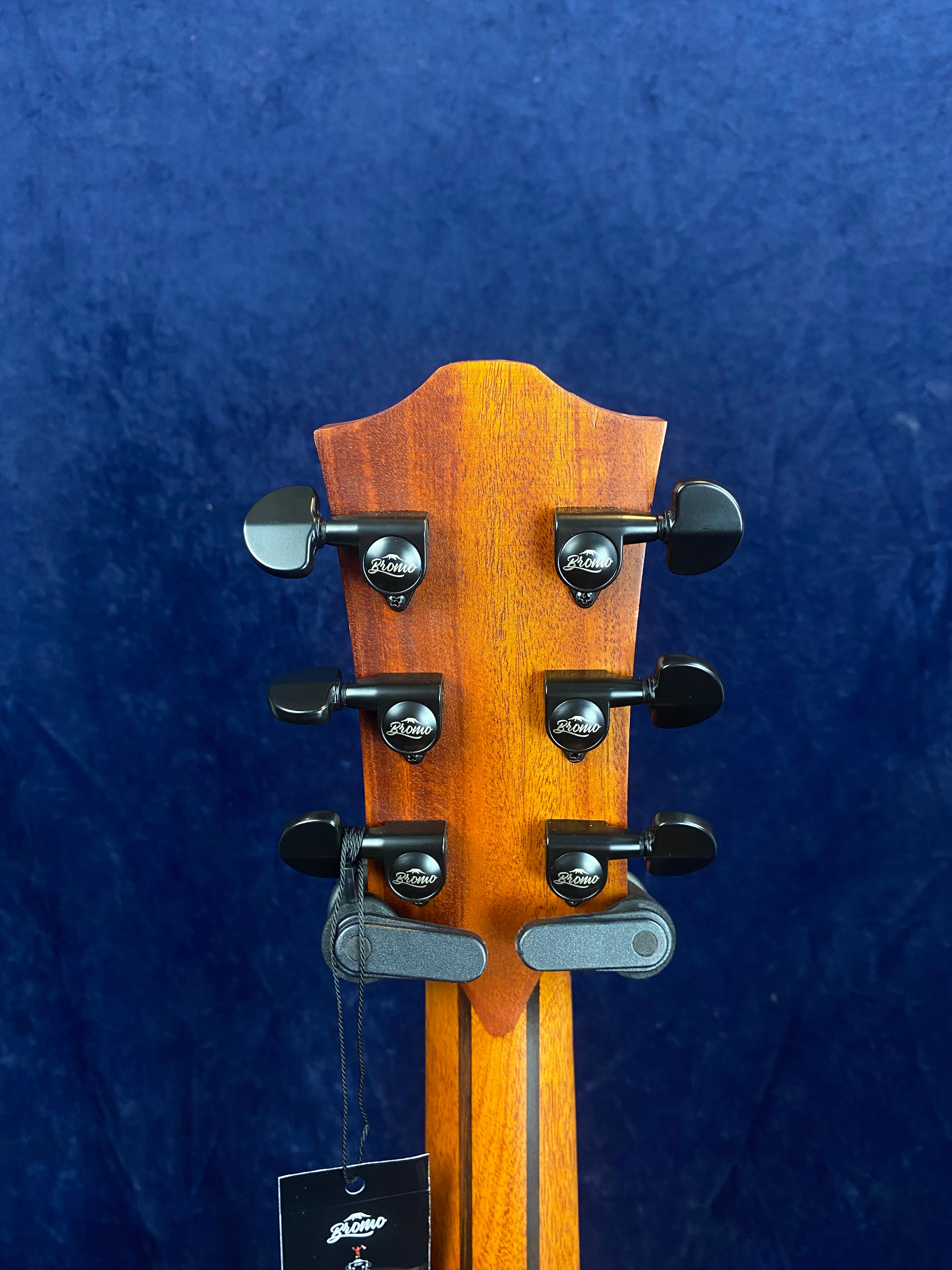 Wood acoustic guitar headstock and neck, six black tuning pegs labeled "Bromo," against a dark blue background, with a black tag hanging below.