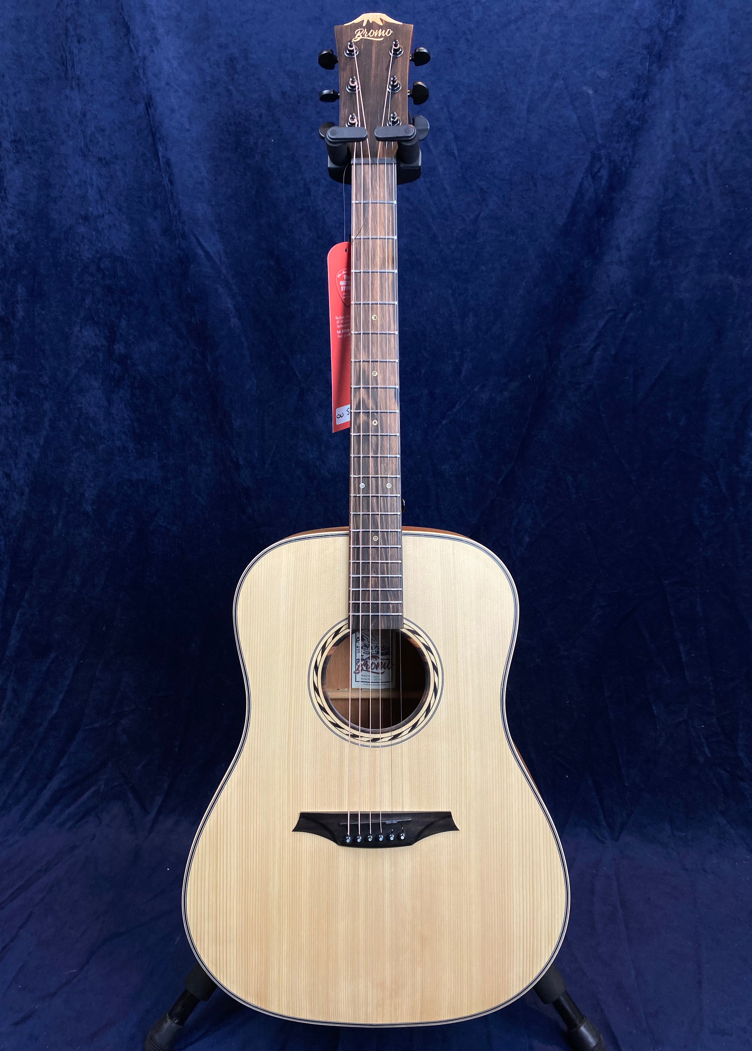 Light wood acoustic guitar with a round sound hole, dark wood bridge and fretboard, black tuning pegs, and a tag hanging from the neck.