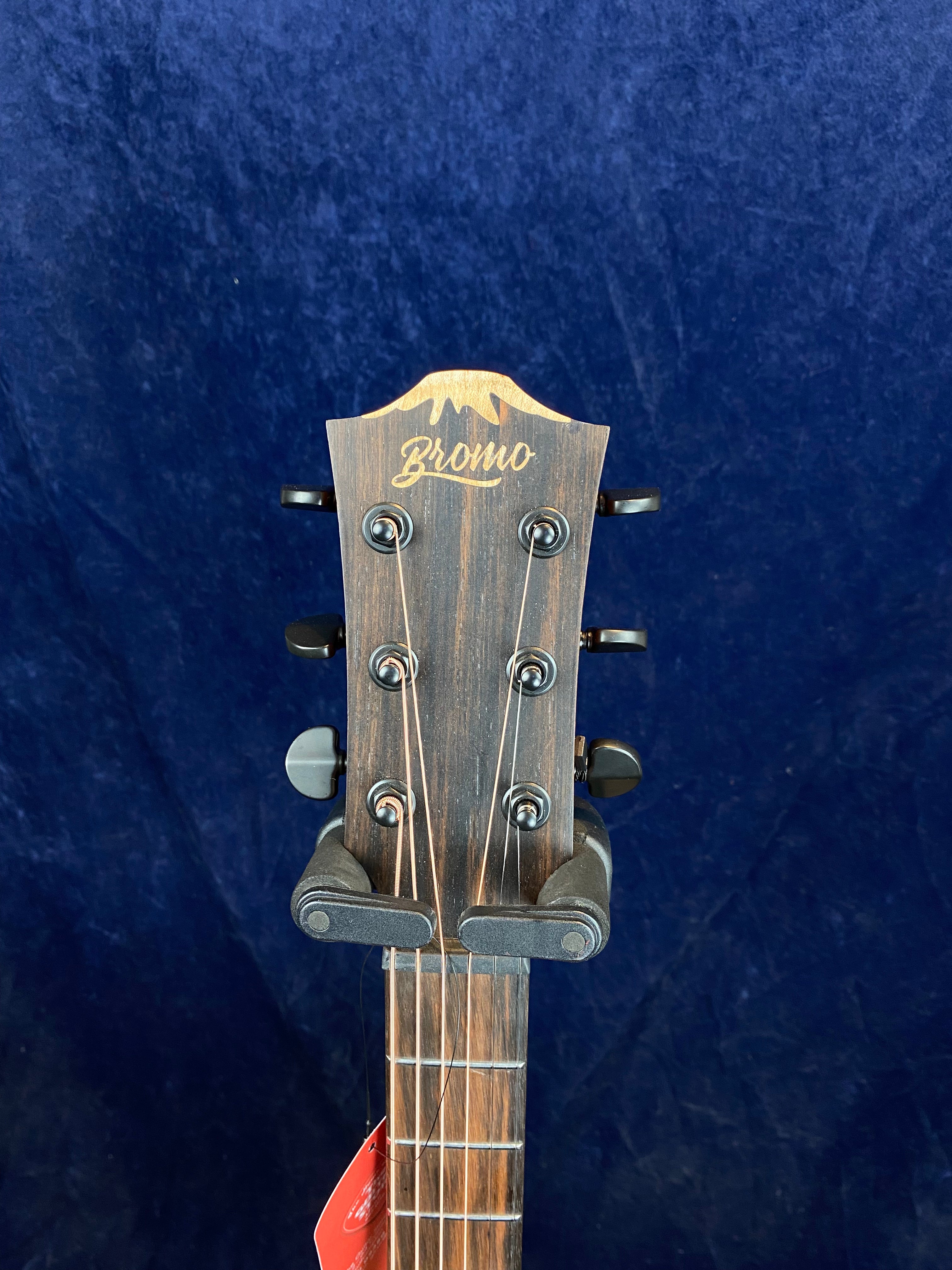 Close-up of a Bromo acoustic guitar headstock with six tuning pegs, wood finish, and a visible guitar hanger against a dark blue background.