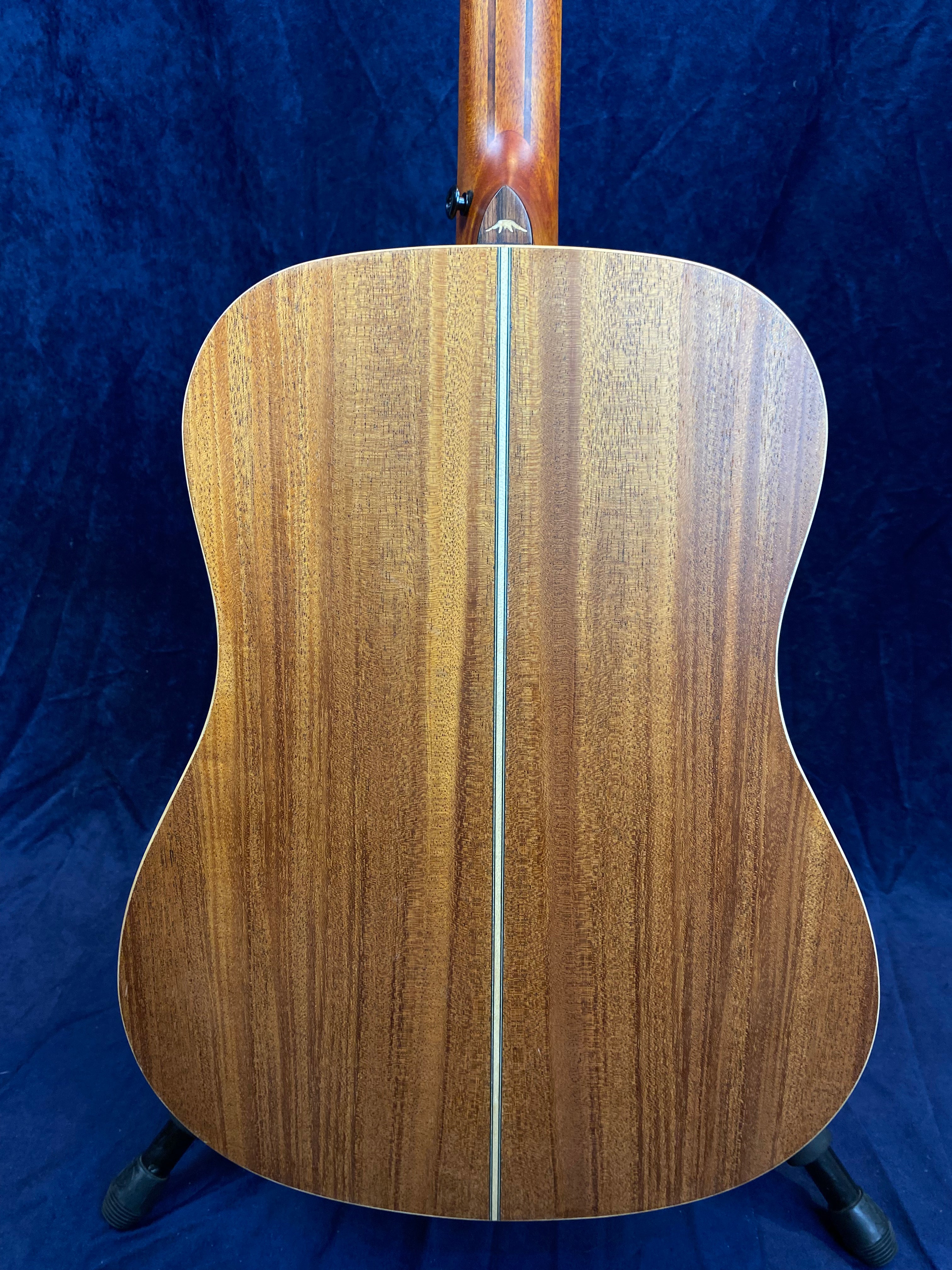 Back of an acoustic guitar in natural wood finish with a thin decorative strip running vertically down the center, set against a dark blue background.