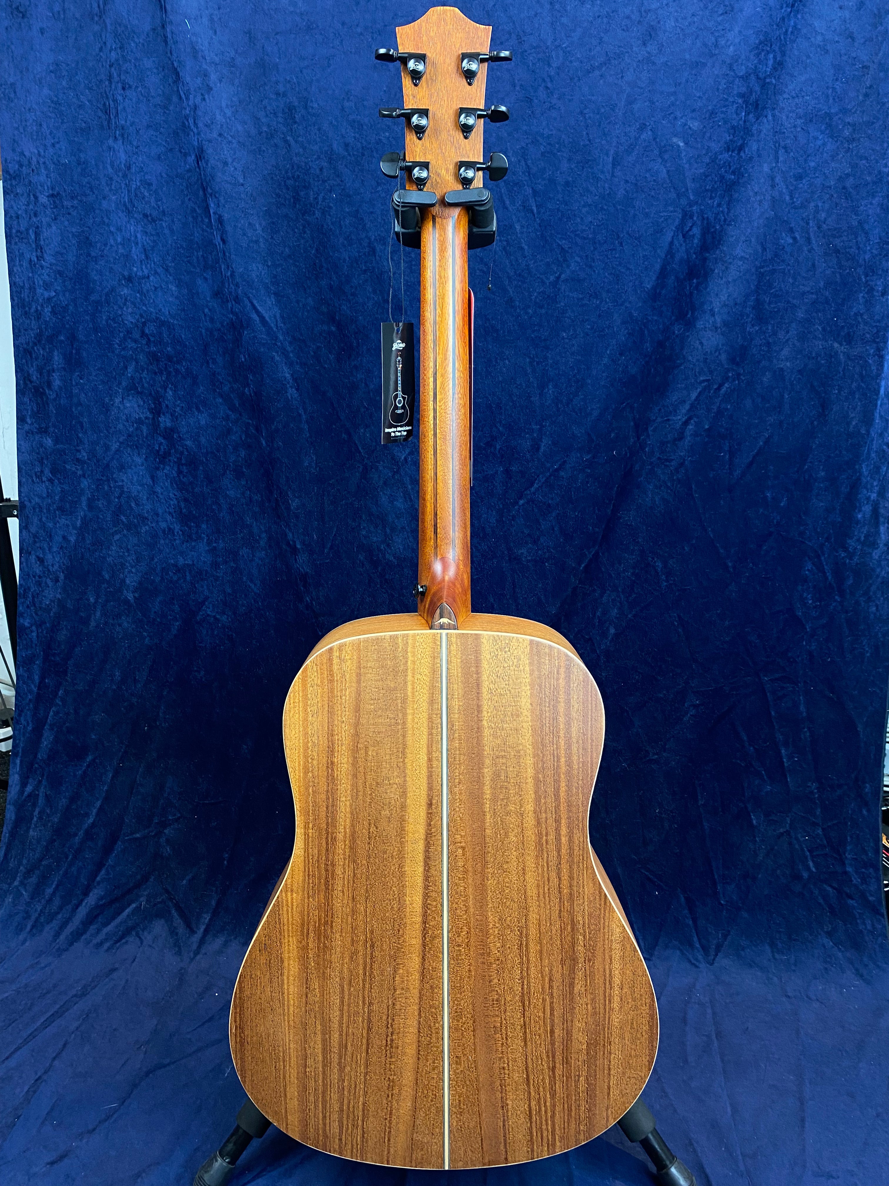 Acoustic guitar shown from the back, displaying a natural wood finish with a central inlay strip and six tuning pegs on the headstock.