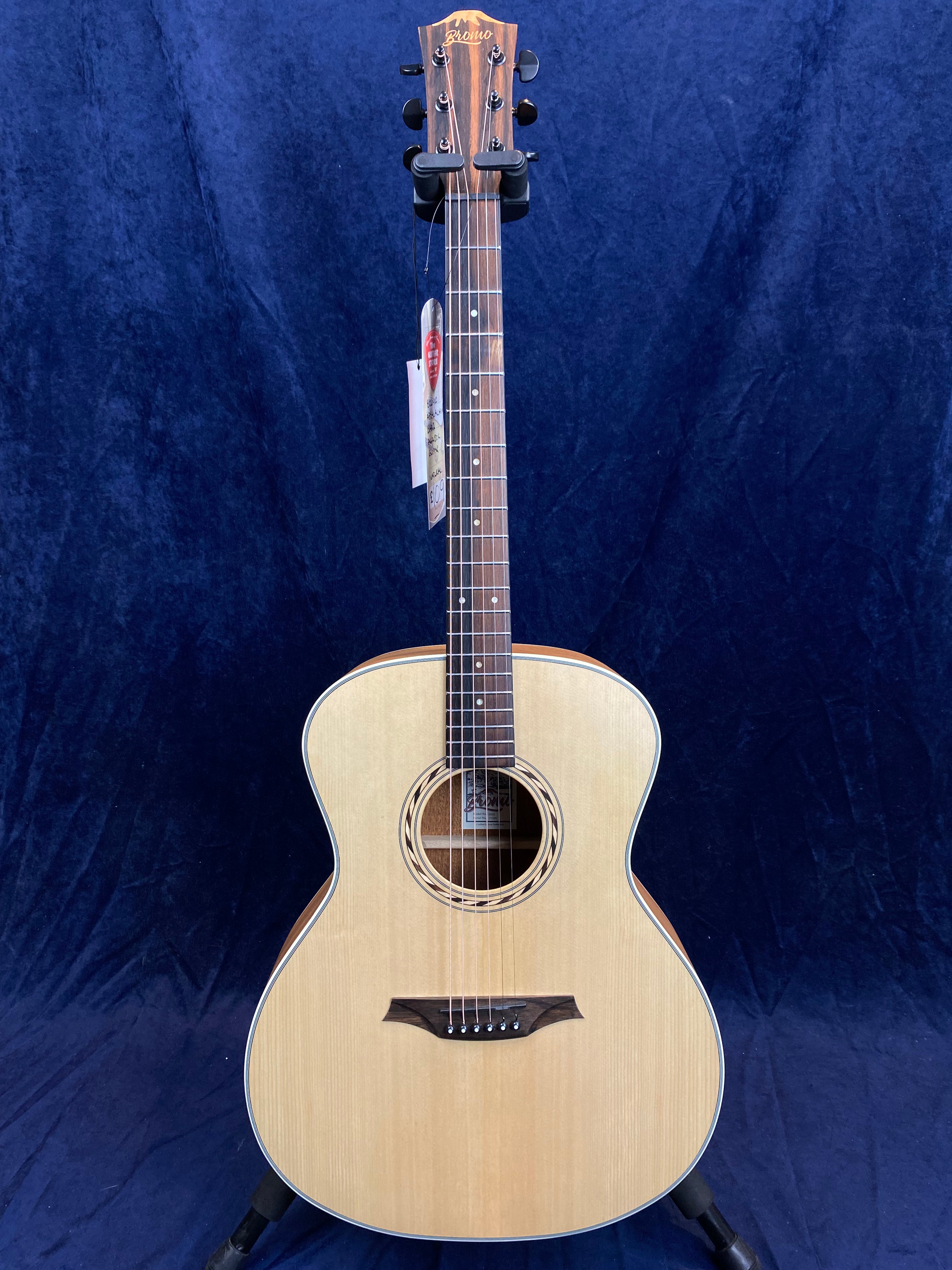 Natural wood acoustic guitar with a light-colored top, dark fretboard, circular sound hole, and six tuning pegs, displayed upright against a dark background.