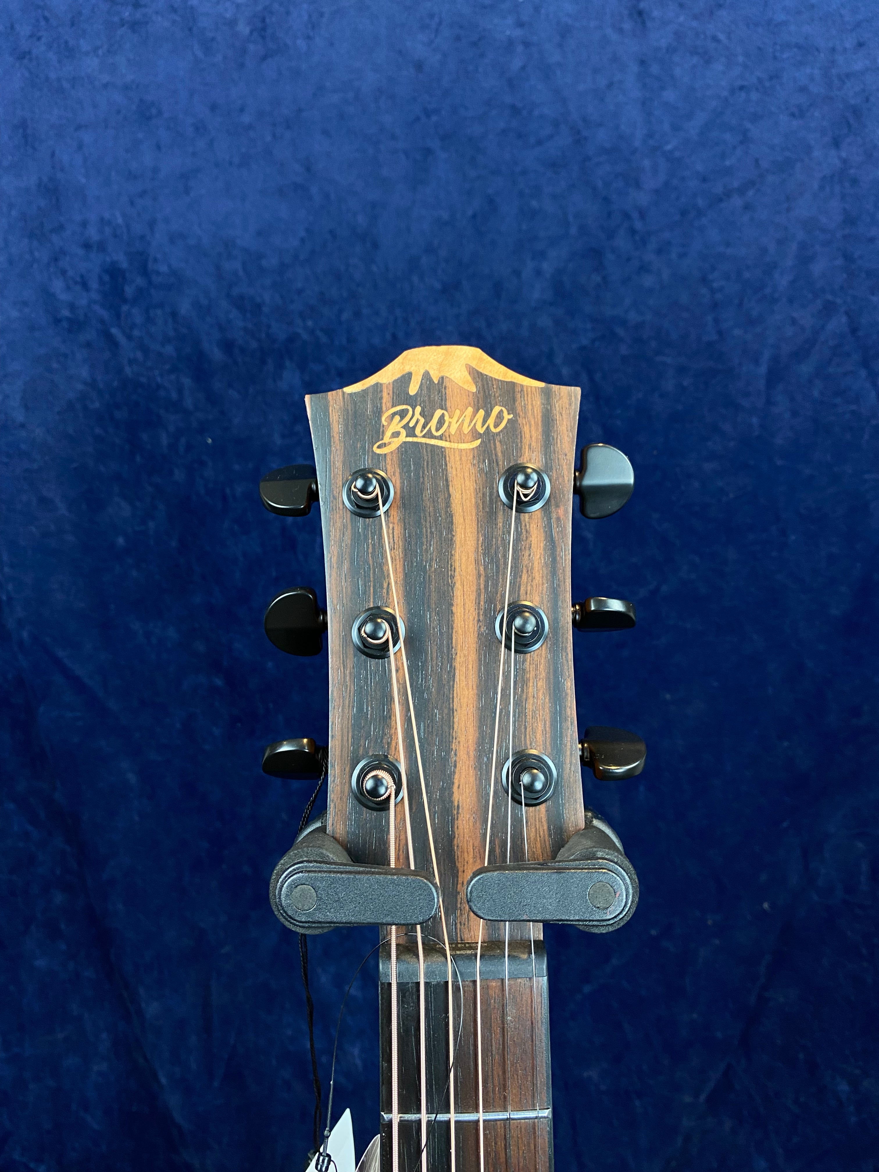 The headstock of a Bromo acoustic guitar with six tuning pegs, natural wood grain, and strings, set against a dark blue background.