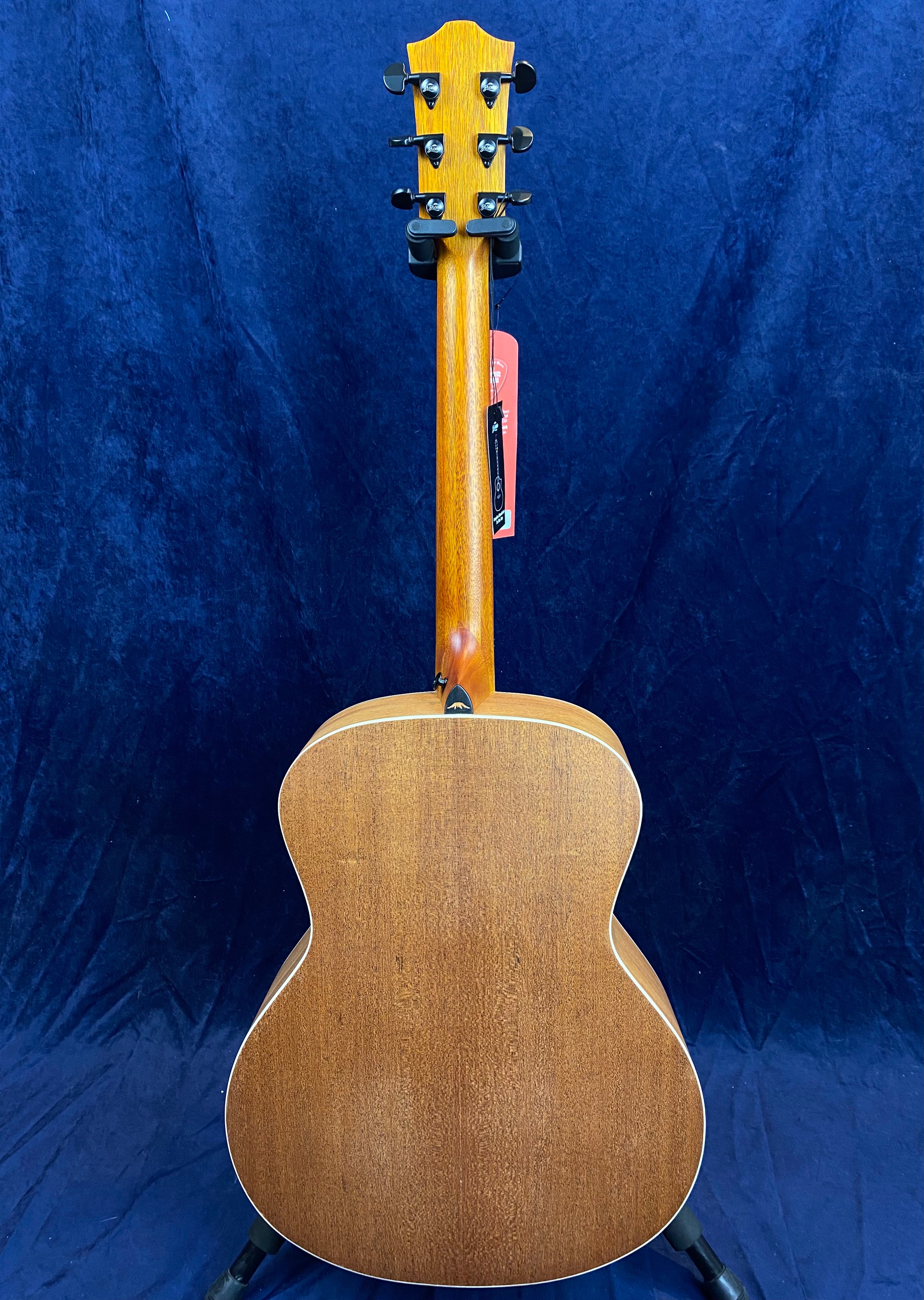 Back view of an acoustic guitar with a natural wood finish, visible tuning pegs, and a tag hanging from the headstock against a dark blue background.