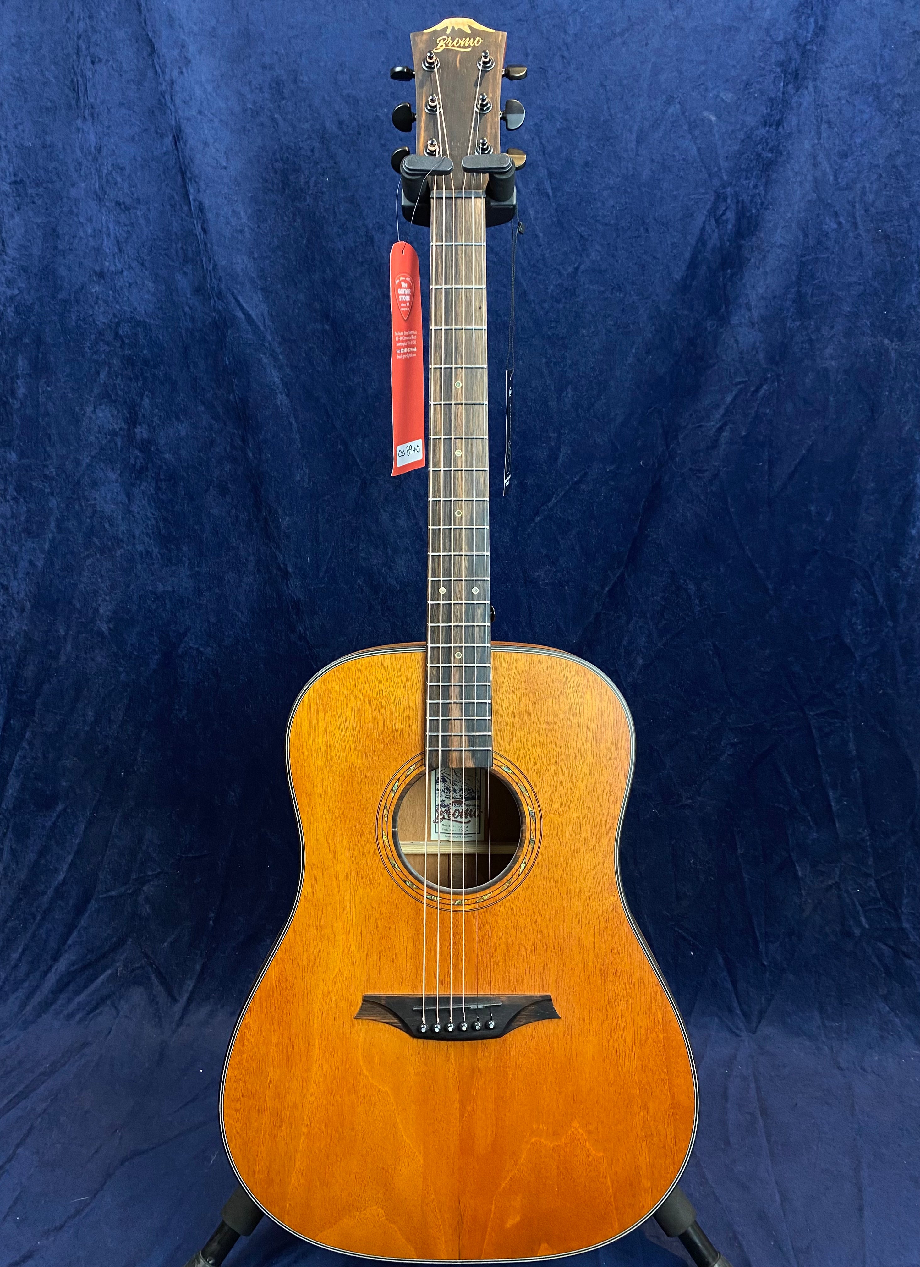 Light brown acoustic guitar with a glossy finish, black tuning pegs, and a red tag hanging from the headstock against a dark blue background.