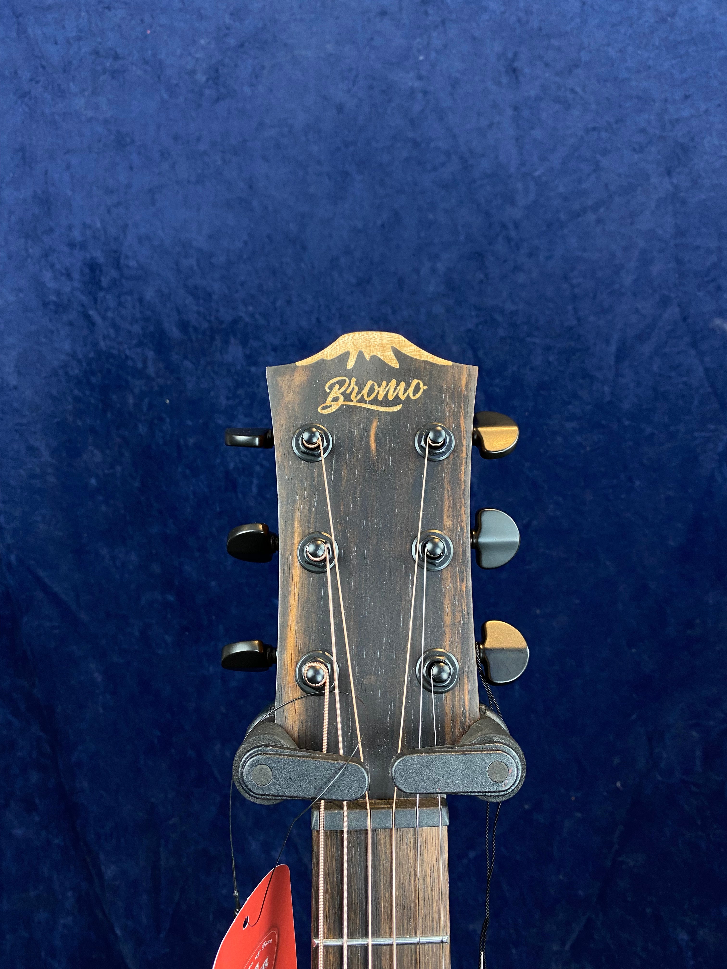 The headstock of a Bromo acoustic guitar, showing six tuning pegs and metal strings against a dark blue background.