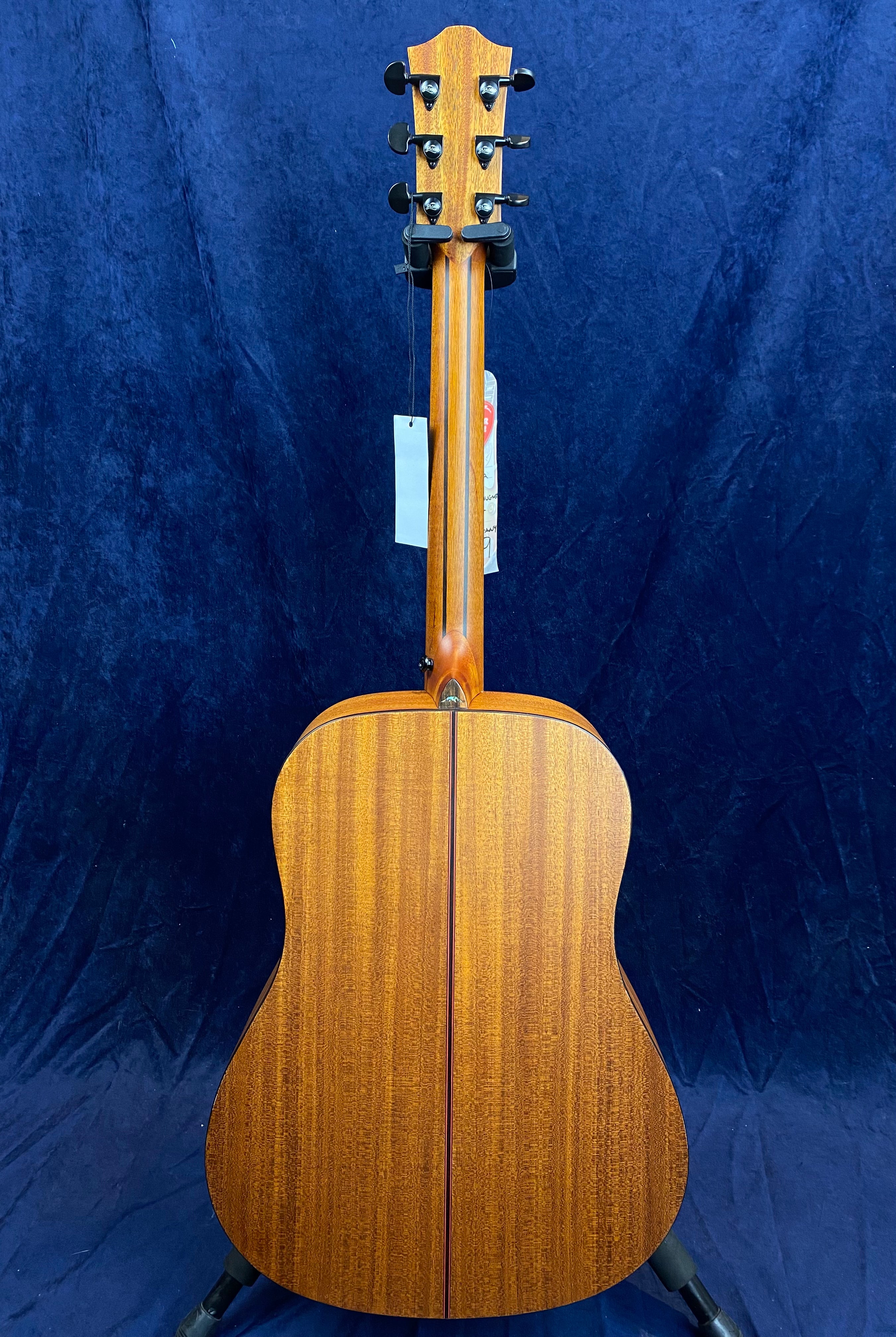 Brown acoustic guitar shown from the back, displaying wood grain, black tuning pegs, and a tag hanging from the neck.
