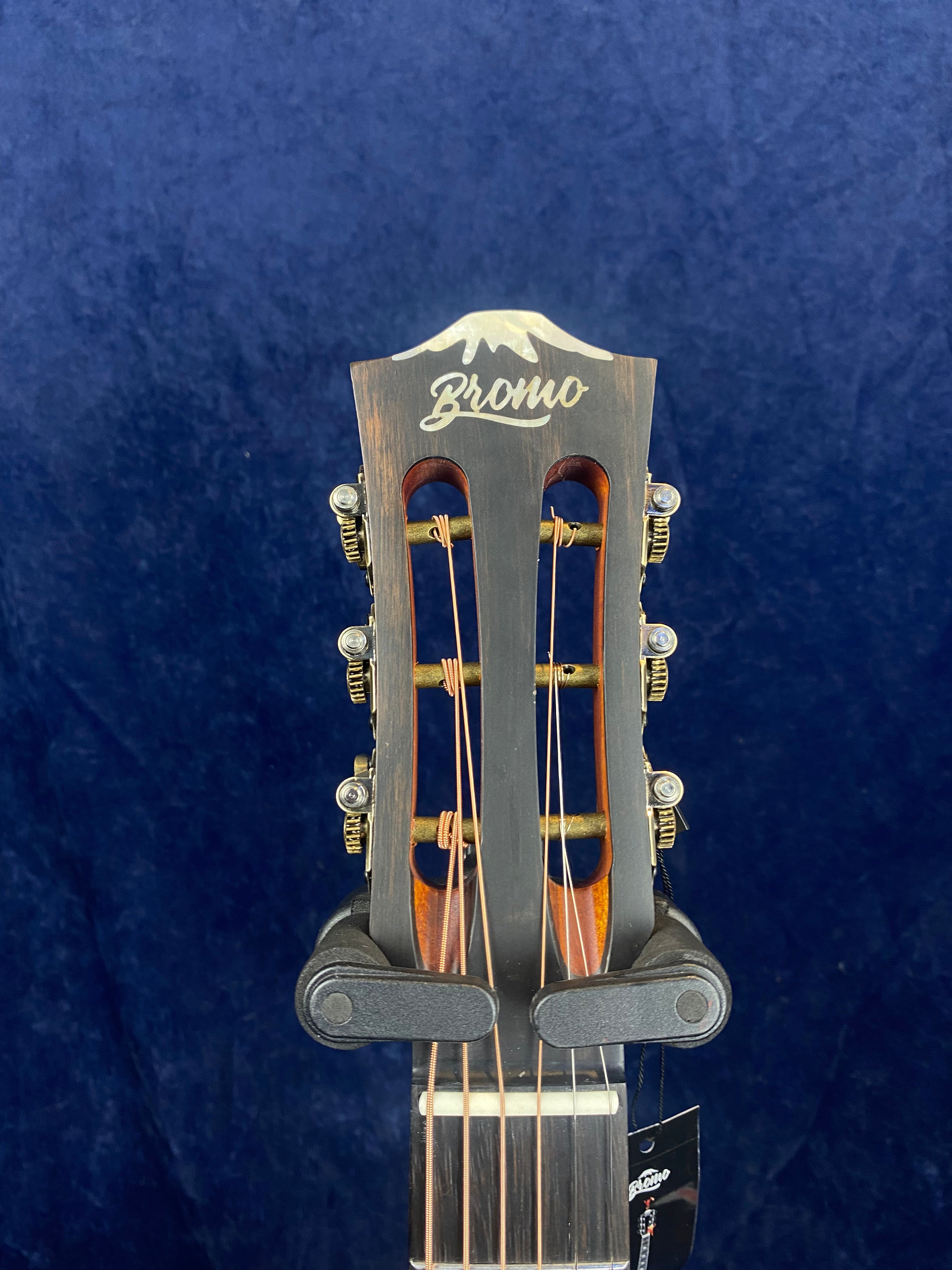 Close-up of a Bromo acoustic guitar headstock with tuning pegs and strings, set against a dark blue background.