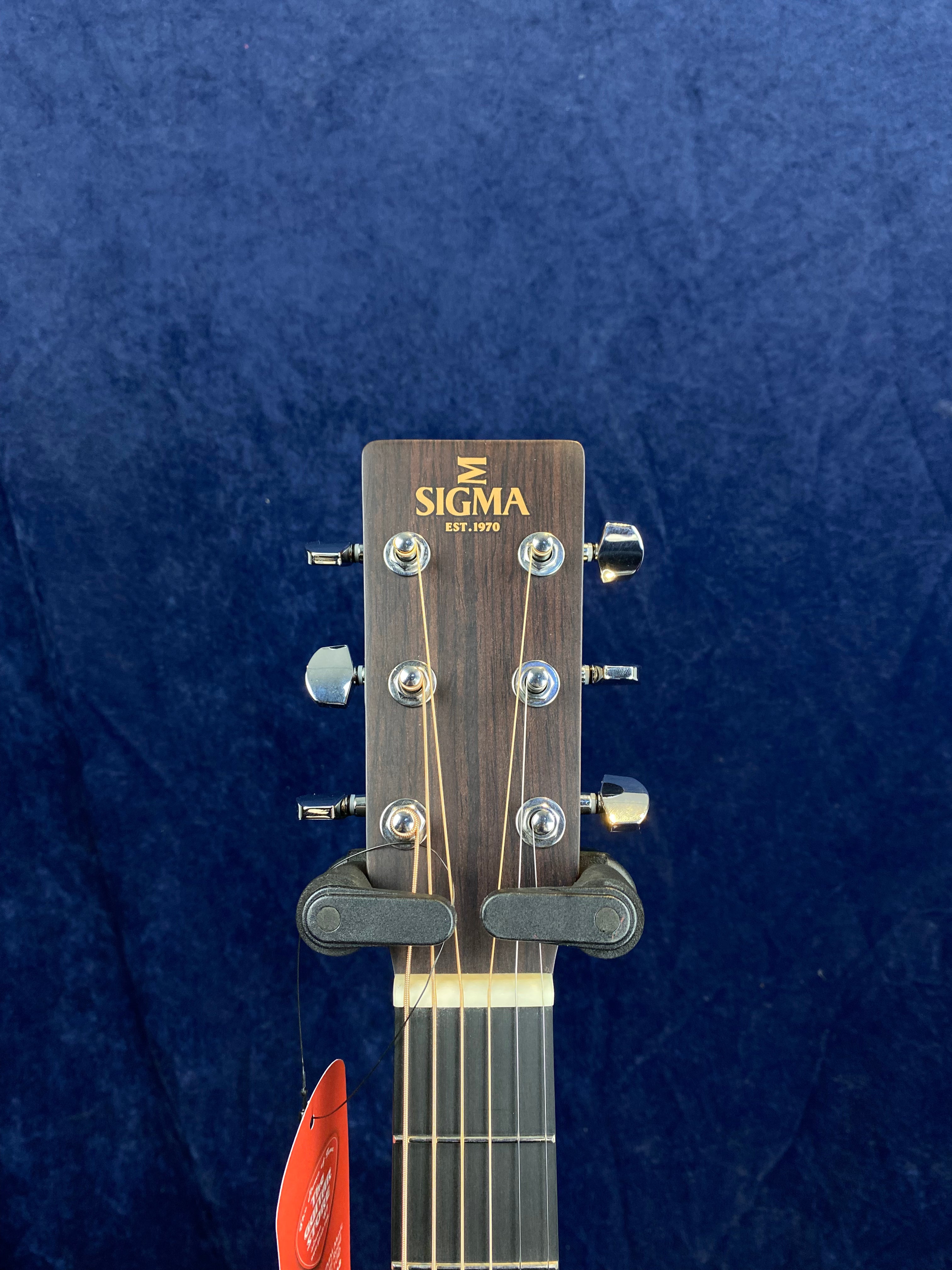 Headstock of a Sigma acoustic guitar with six tuning pegs, wood grain finish, and Sigma logo reading "SIGMA EST. 1970" on dark blue background.