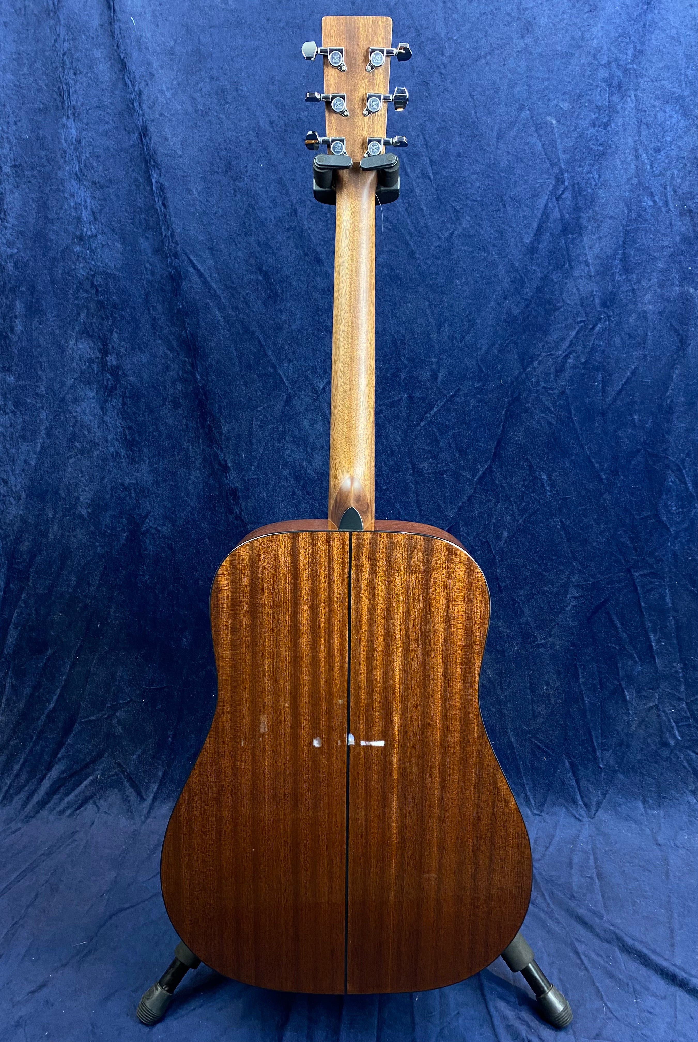 Rear view of an acoustic guitar with a dark wood finish, visible grain, and silver tuning pegs, standing on a black floor stand against a blue backdrop.