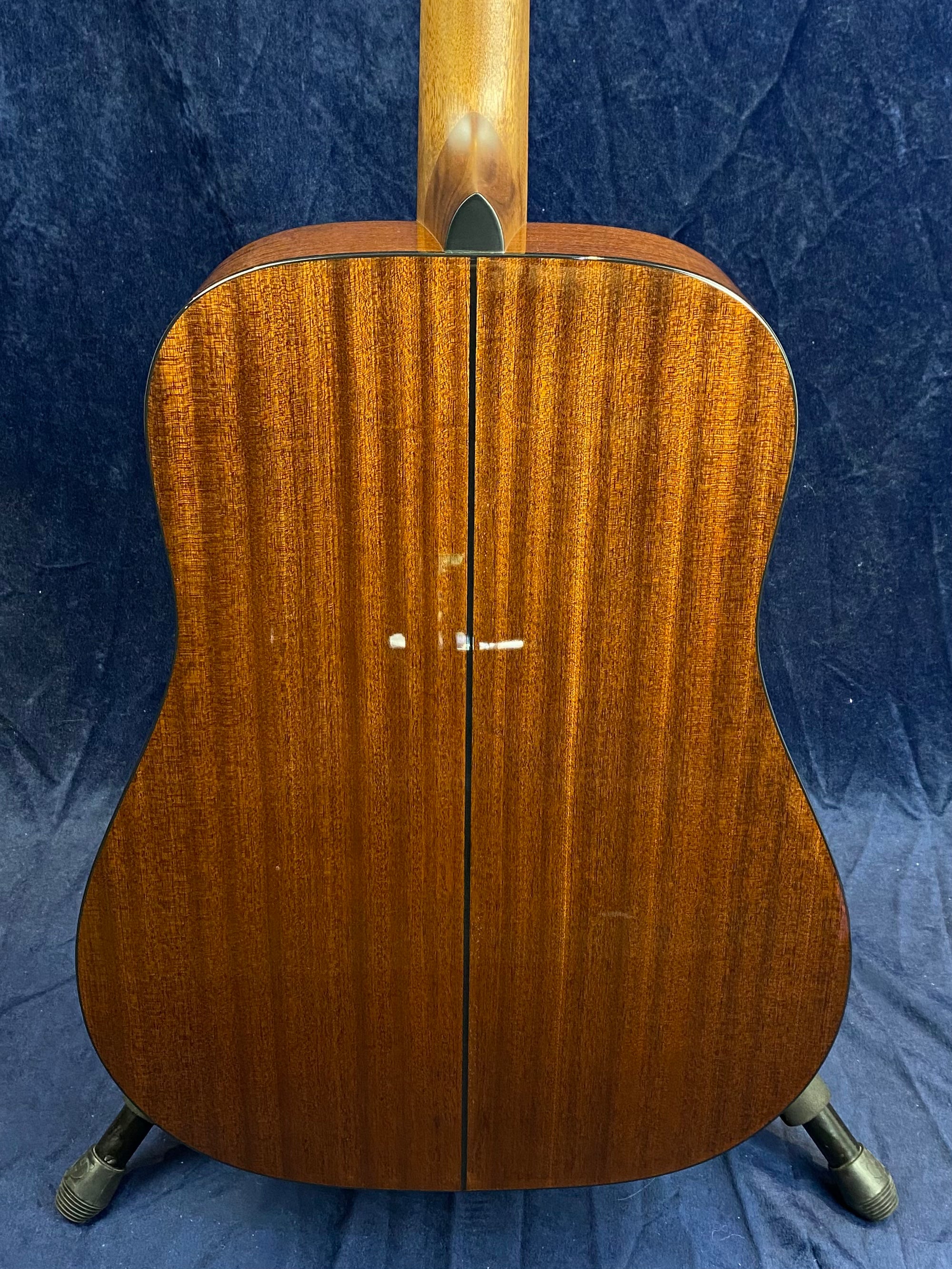 Back view of an acoustic guitar with a rich, dark wood grain finish and a visible center seam, resting on a stand against a dark background.