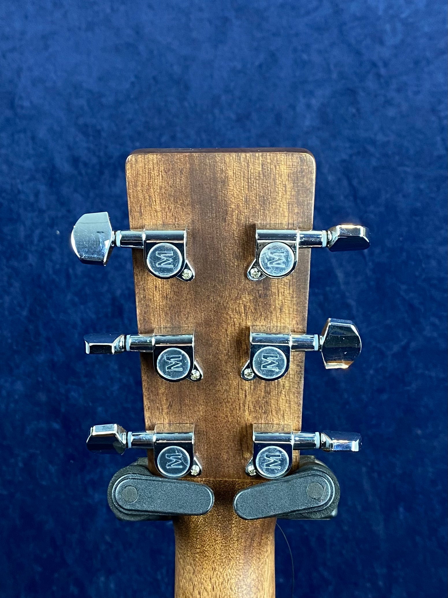 Rear view of an acoustic guitar headstock with six chrome tuning pegs, each marked with a "W" in a circle, against a dark blue background.