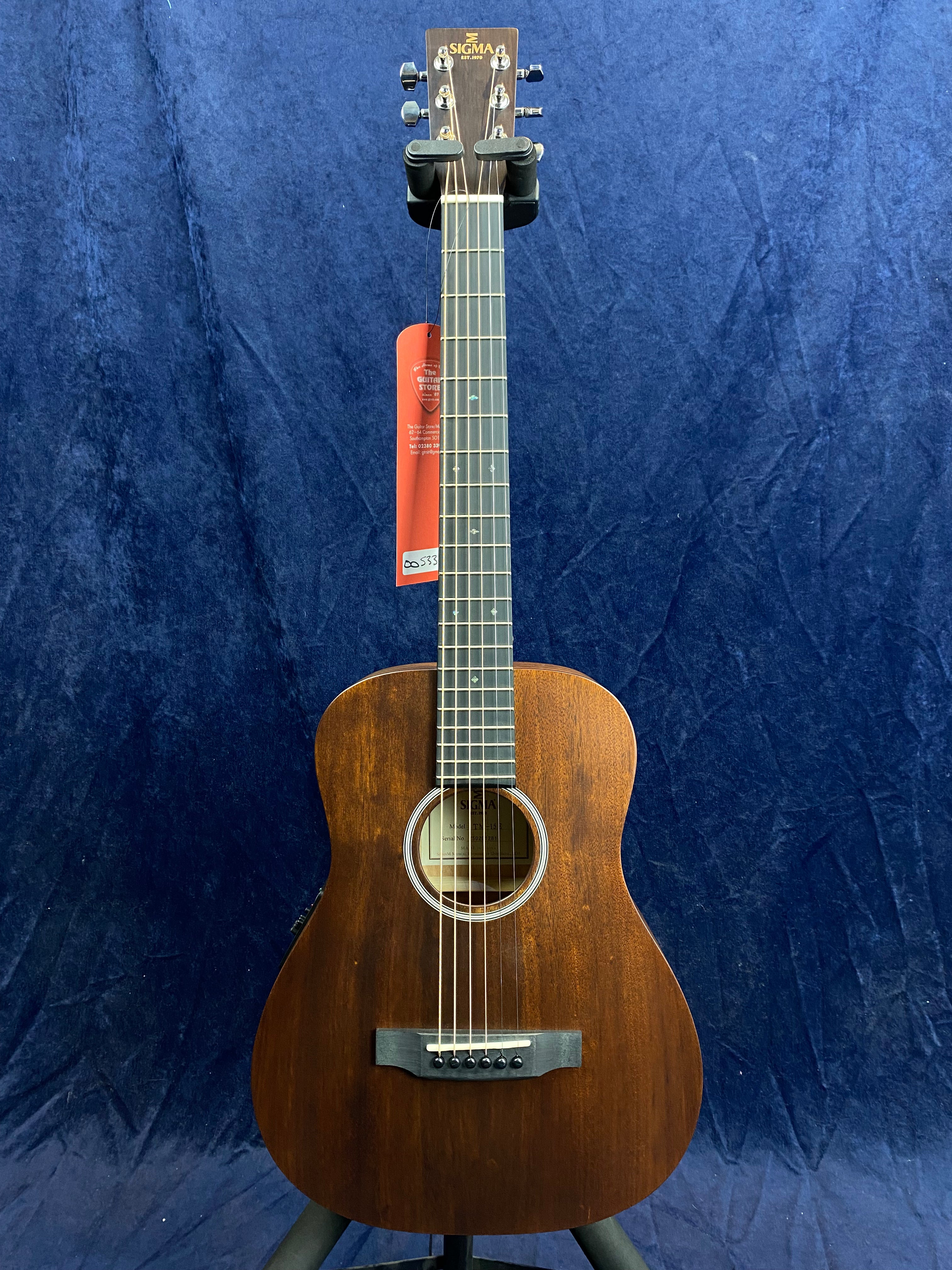 A Sigma acoustic guitar with a dark wood finish, six strings, rounded body, and a central sound hole, shown upright against a dark blue background.