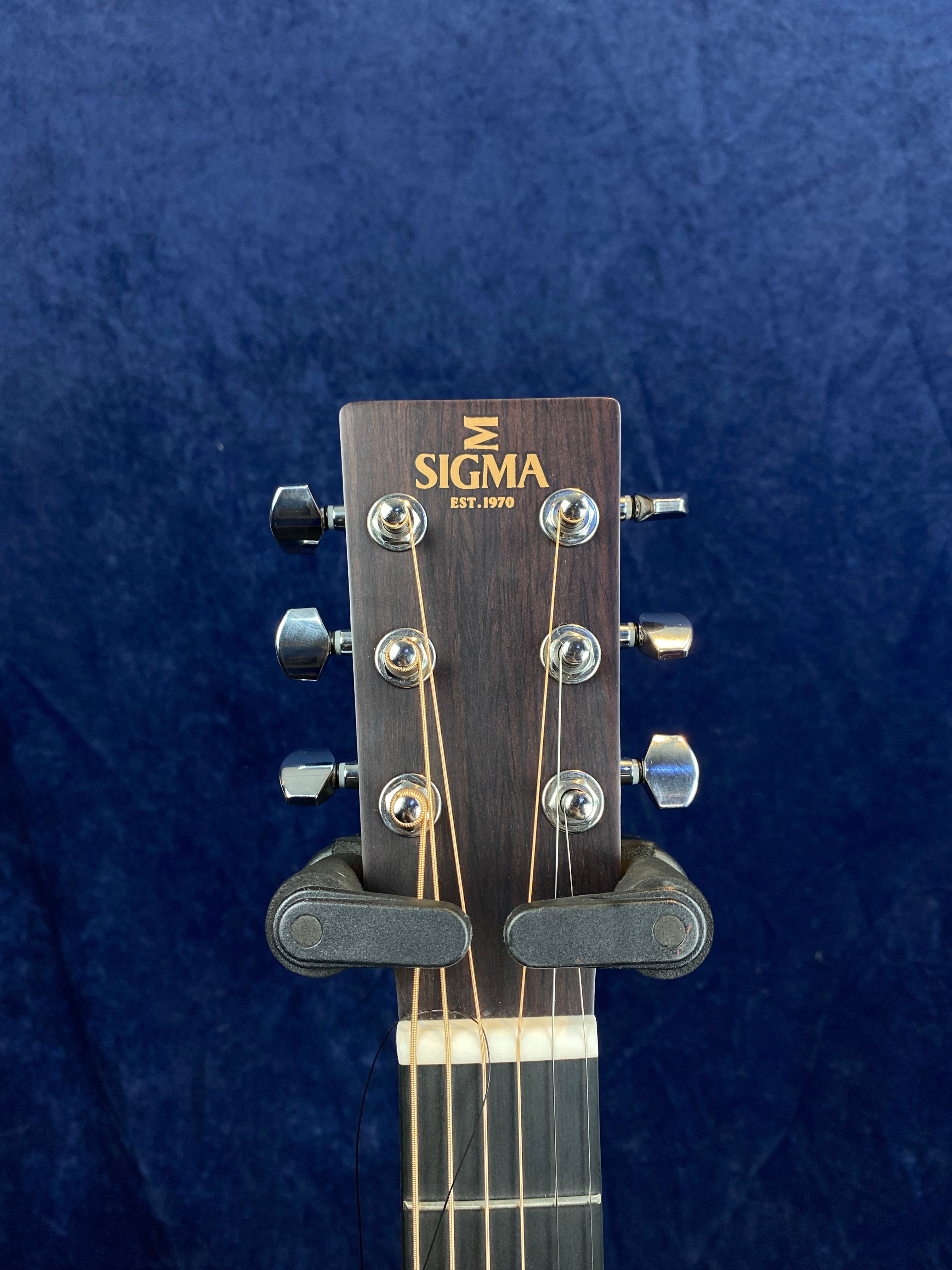 The headstock of a Sigma guitar showing six tuning pegs, logo with "SIGMA EST. 1970," and a capo clamped above the nut on the fretboard.