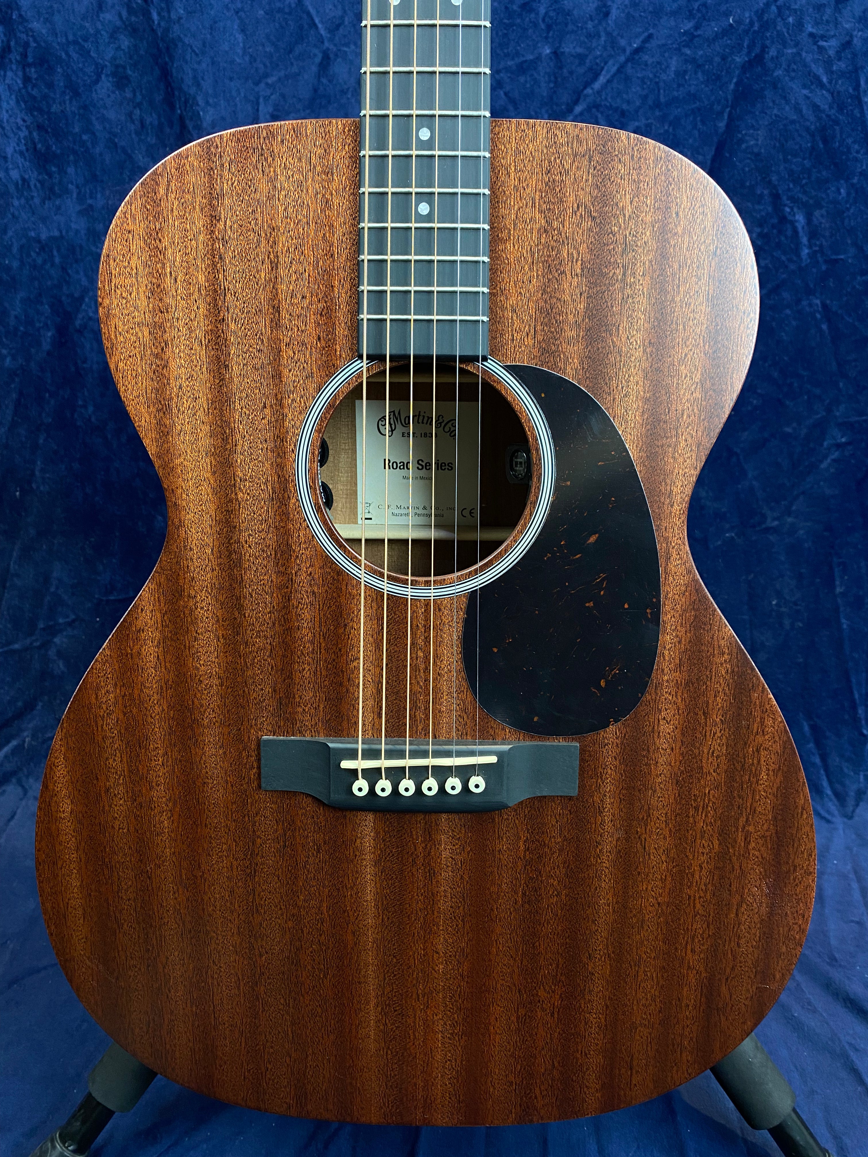 Brown acoustic guitar with a dark wood grain finish, six strings, black pickguard, and white bridge pins set against a dark blue background.