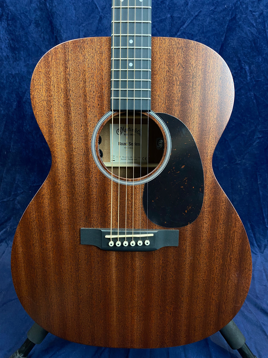 Brown acoustic guitar with a dark wood grain finish, six strings, black pickguard, and white bridge pins set against a dark blue background.