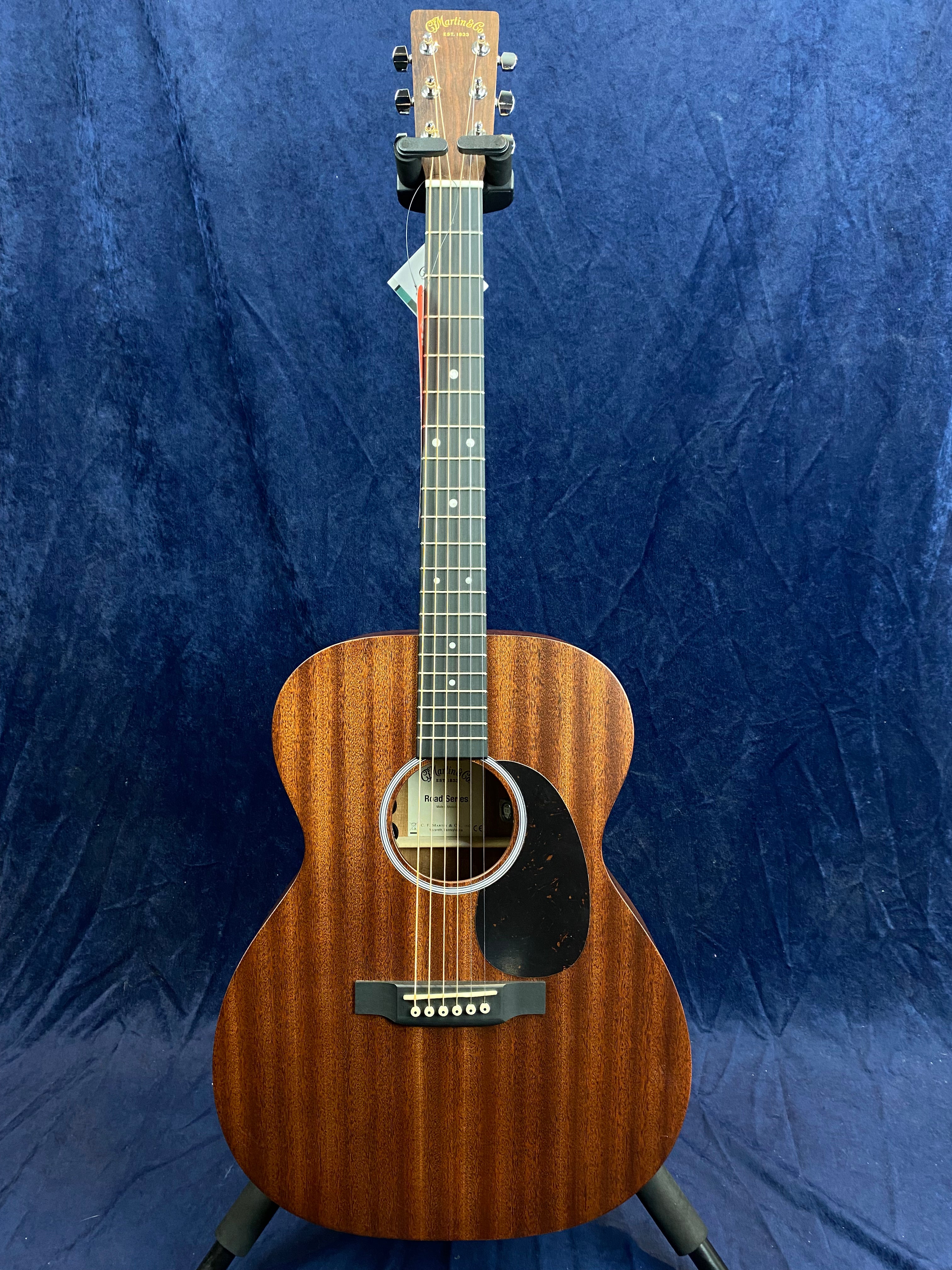 Acoustic guitar with a dark wood finish, six strings, circular sound hole, black pickguard, and white dot fret markers, standing on a stand.