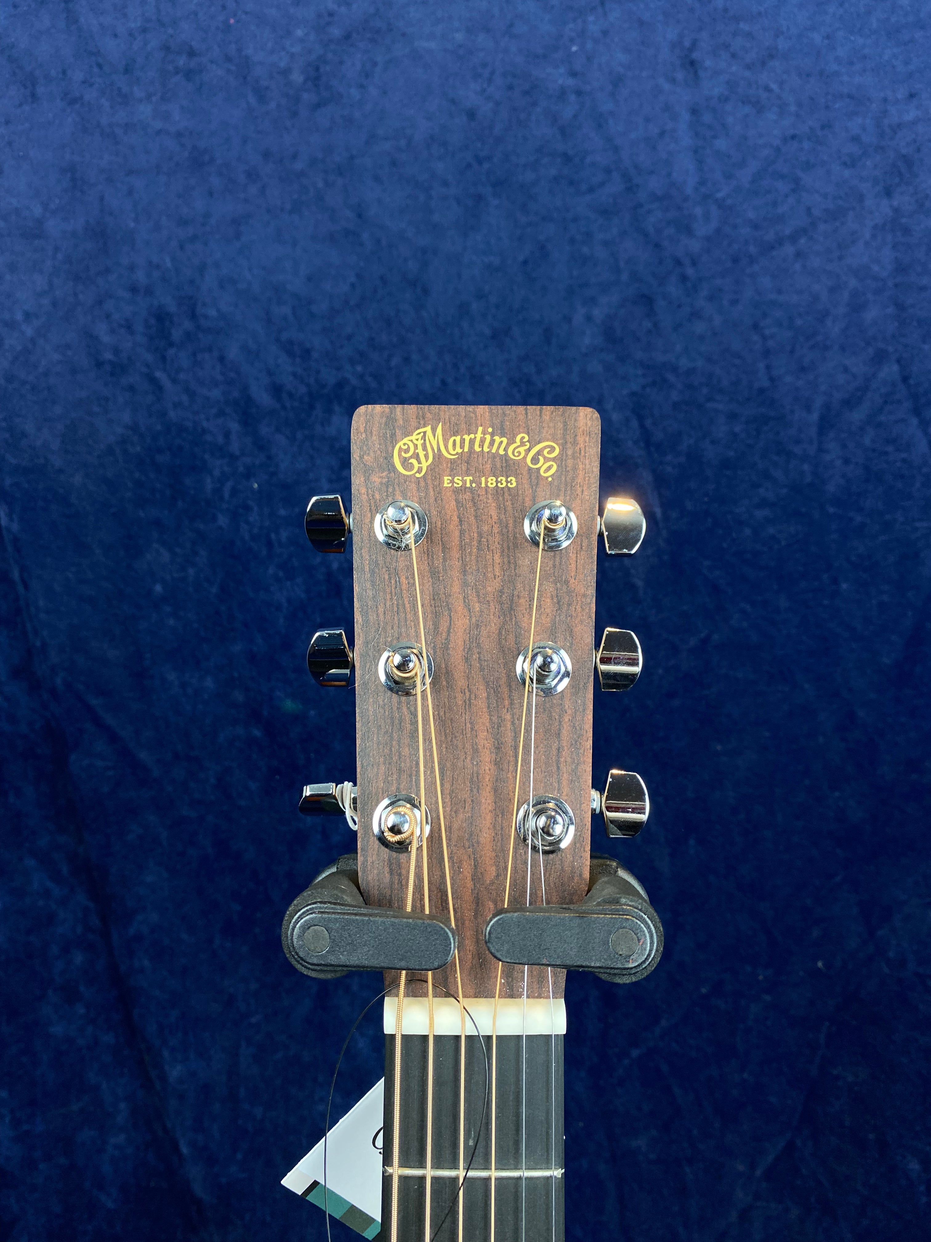 Wood headstock of a Martin & Co. acoustic guitar with six silver tuning pegs, cream nut, and gold logo text, against a dark blue background.