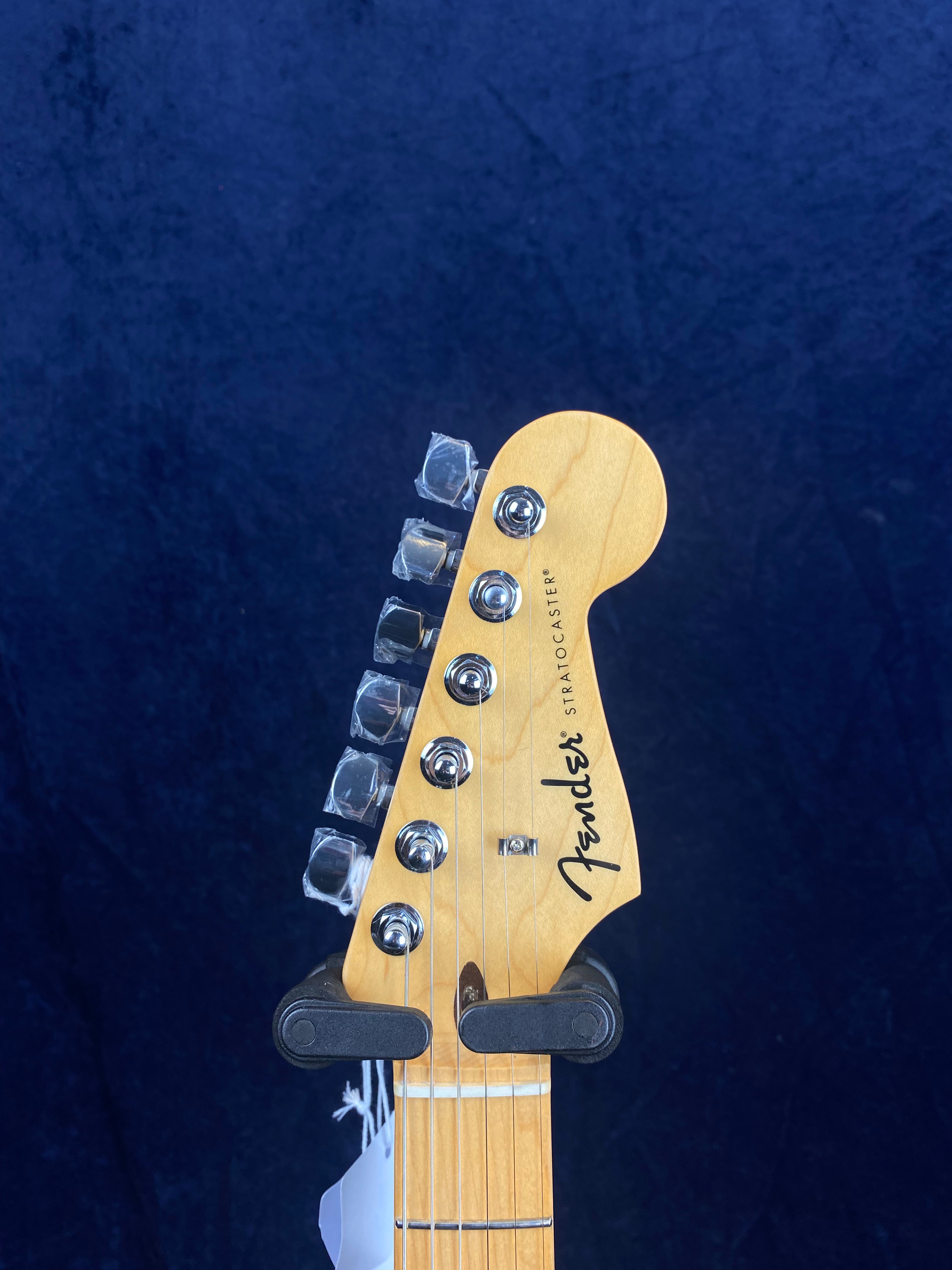 Light wood Fender Stratocaster electric guitar headstock with six tuning pegs, black logo, and part of the neck visible against a dark background.