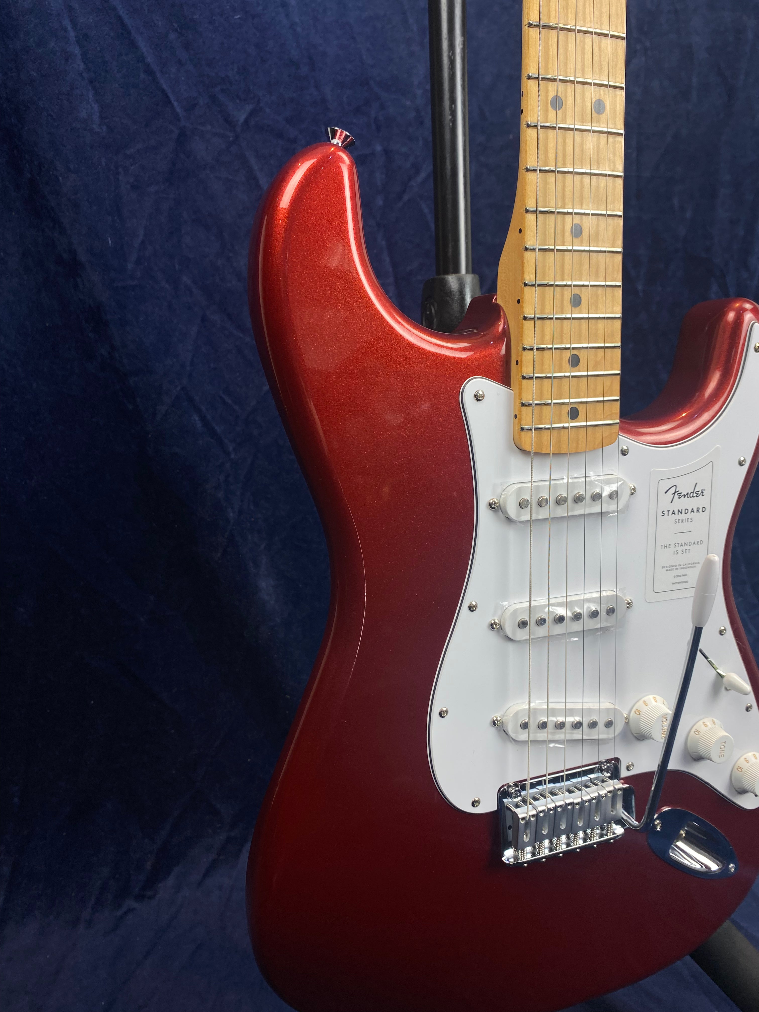 Red Fender electric guitar with a white pickguard, three single-coil pickups, and a maple fretboard, shown on a stand against a dark background.