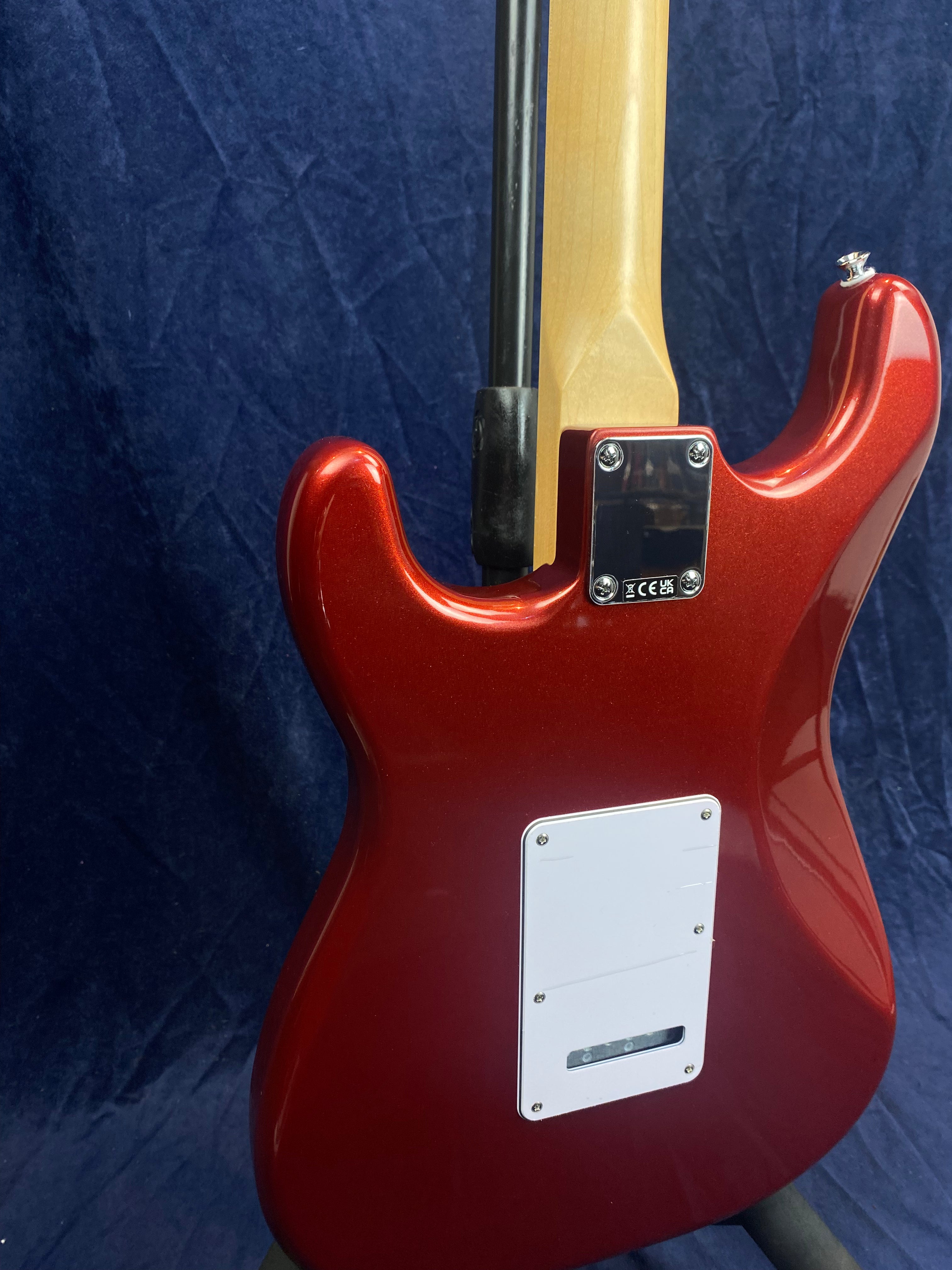 Red electric guitar back with a white cover plate and light wood neck, propped on a stand against a dark blue textured fabric background.