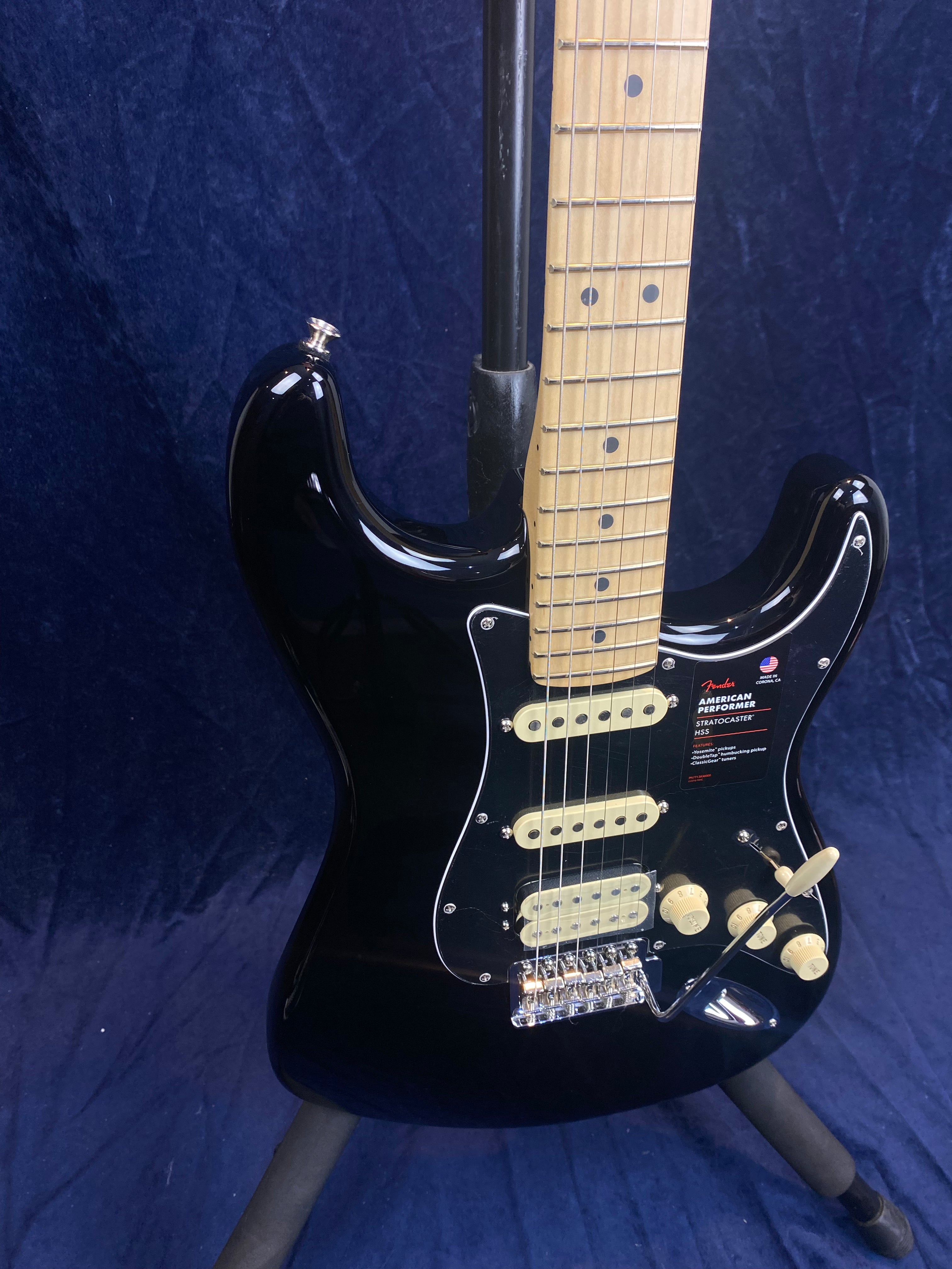 Black Fender electric guitar with a maple neck, cream pickups and knobs, chrome bridge, and a whammy bar, shown on a stand against a dark backdrop.