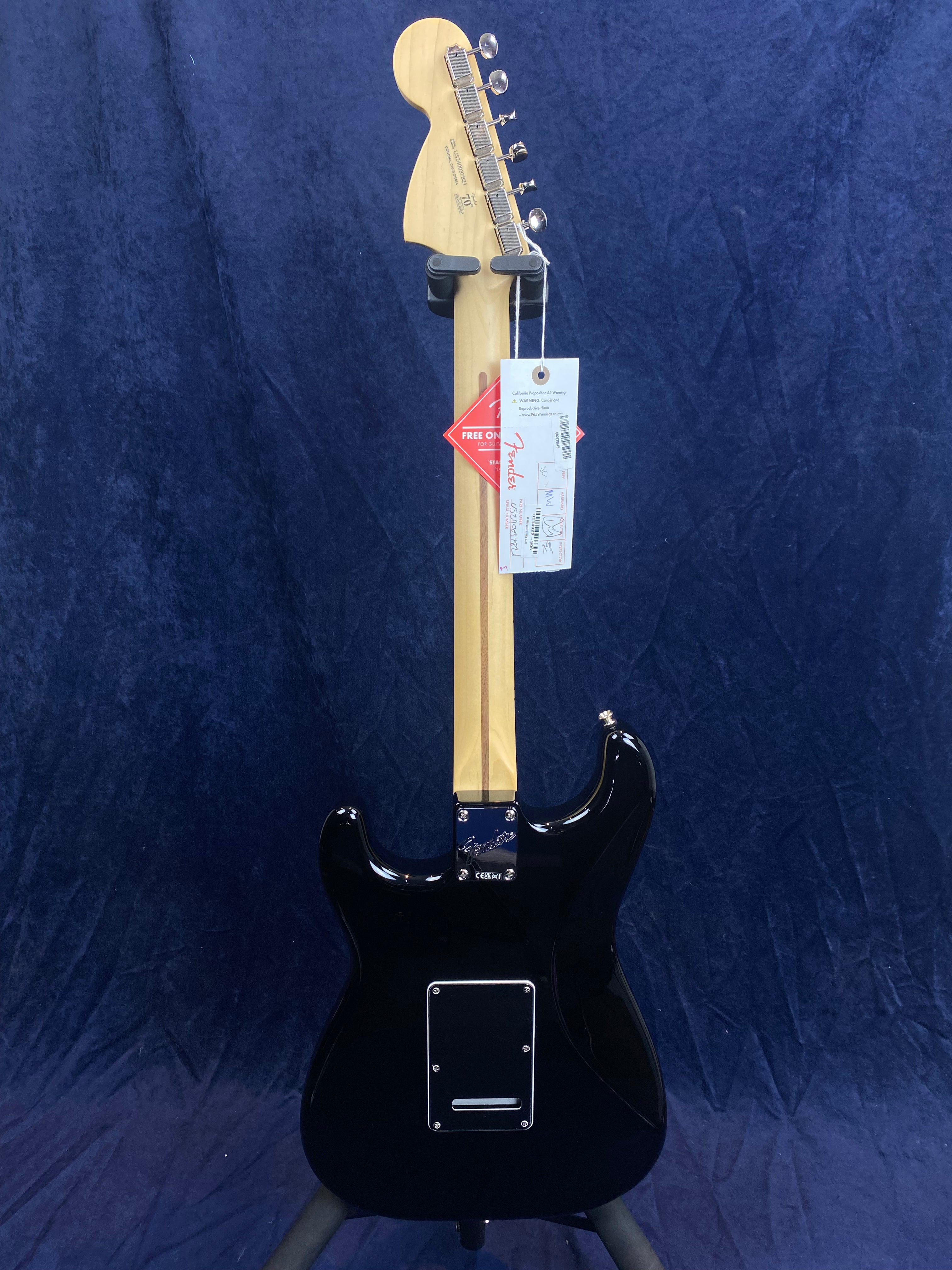 Black electric guitar shown from the back with a light wood neck, metal tuning pegs, and retail tags hanging from the headstock.
