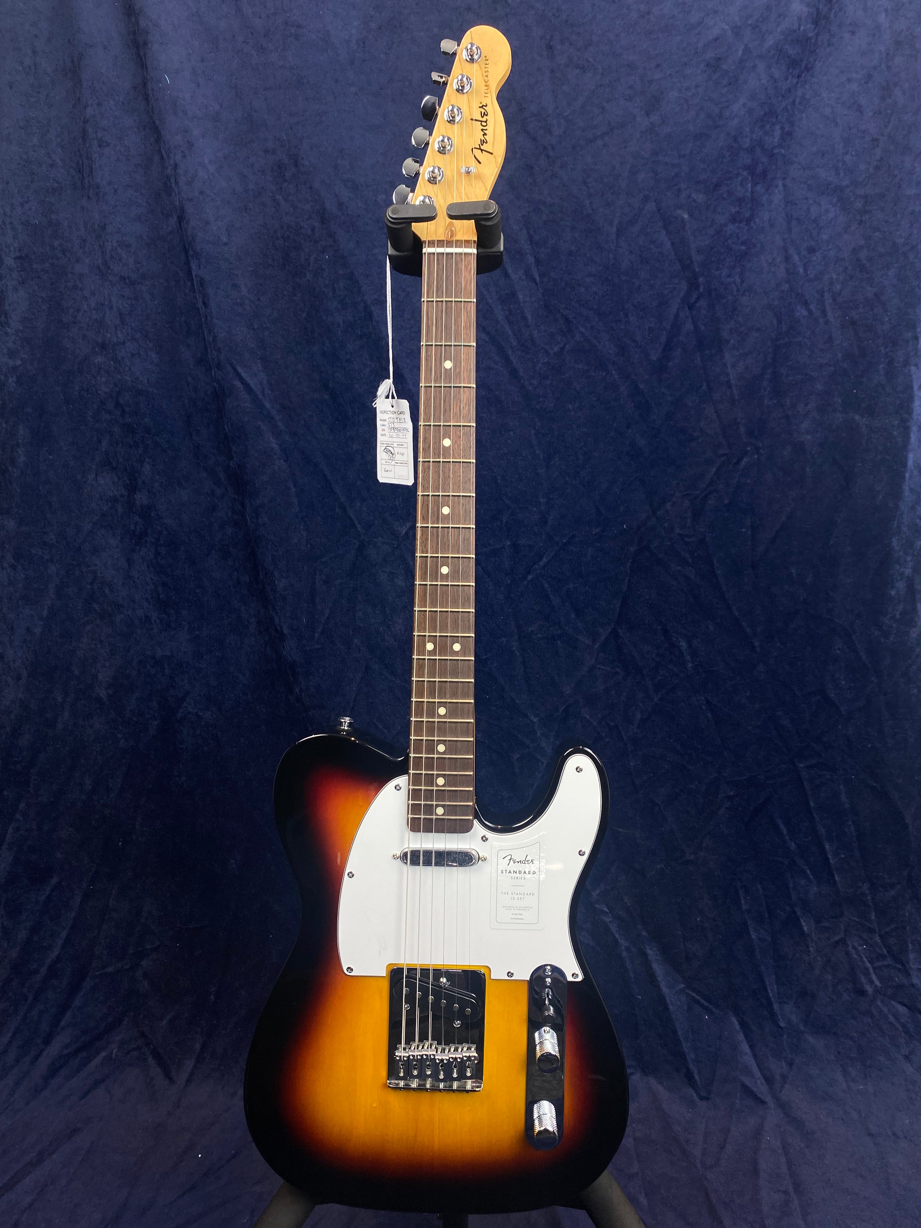 Fender electric guitar with a sunburst body, white pickguard, chrome hardware, and six tuning pegs against a dark blue background.