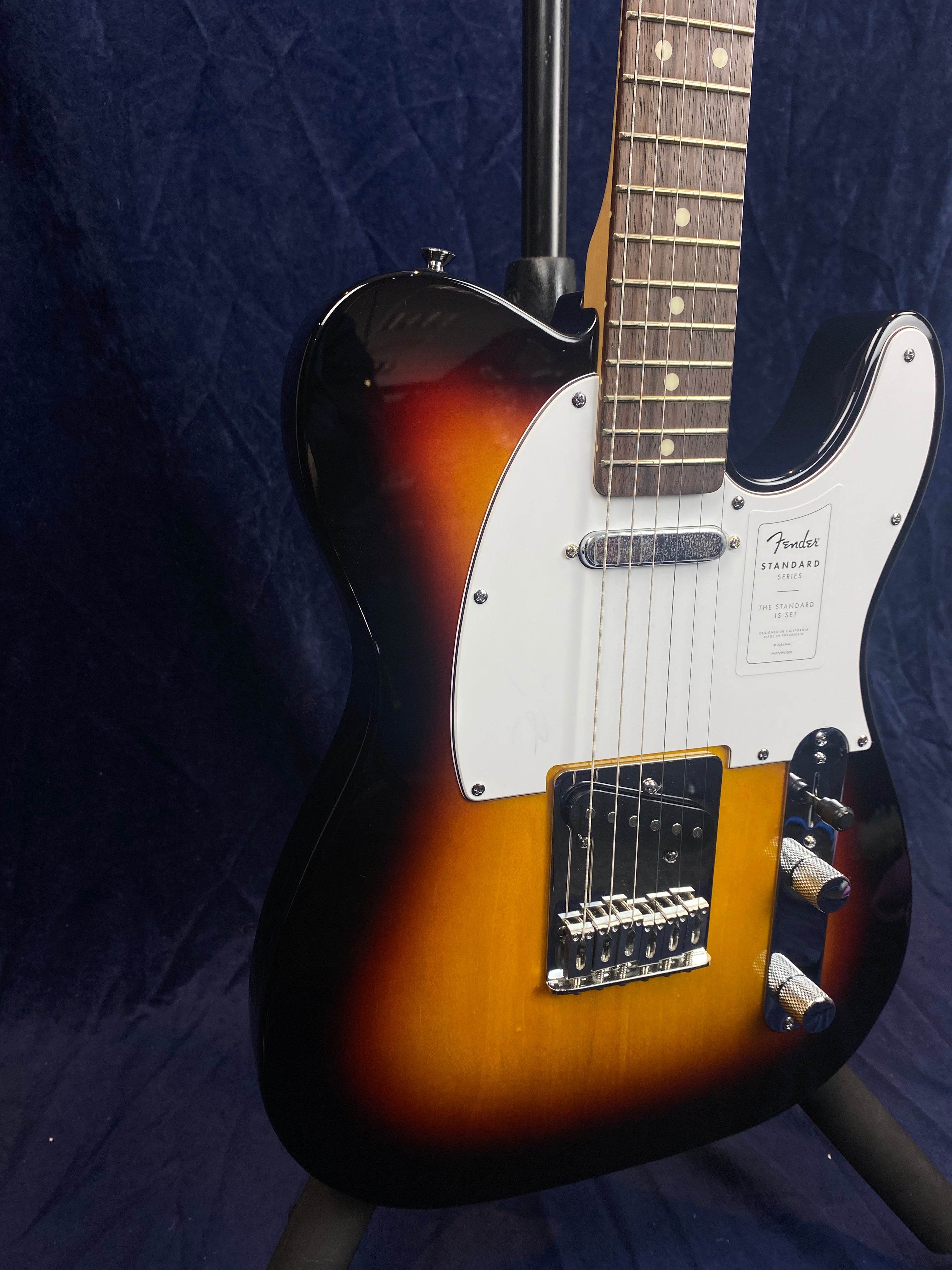 Sunburst Fender electric guitar with a white pickguard, chrome knobs, and a rosewood fretboard, shown on a stand against a dark background.