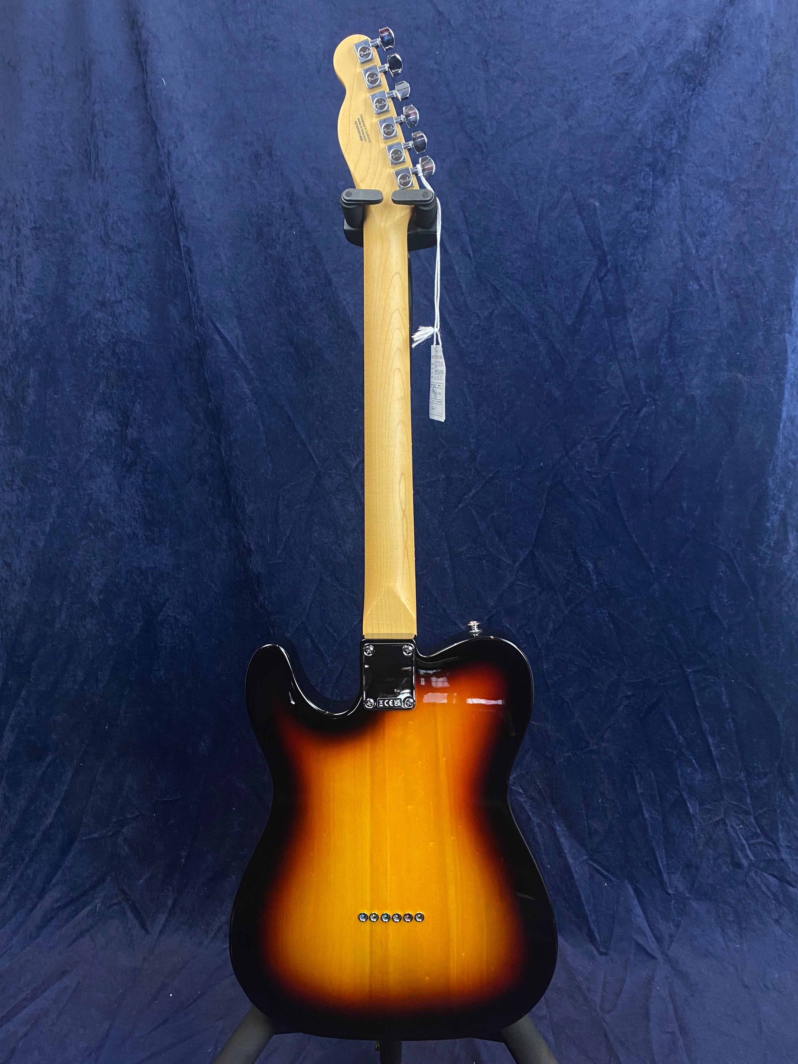 Sunburst electric guitar shown from the back, with a maple neck and six tuning pegs, hanging on a stand against a dark blue background.