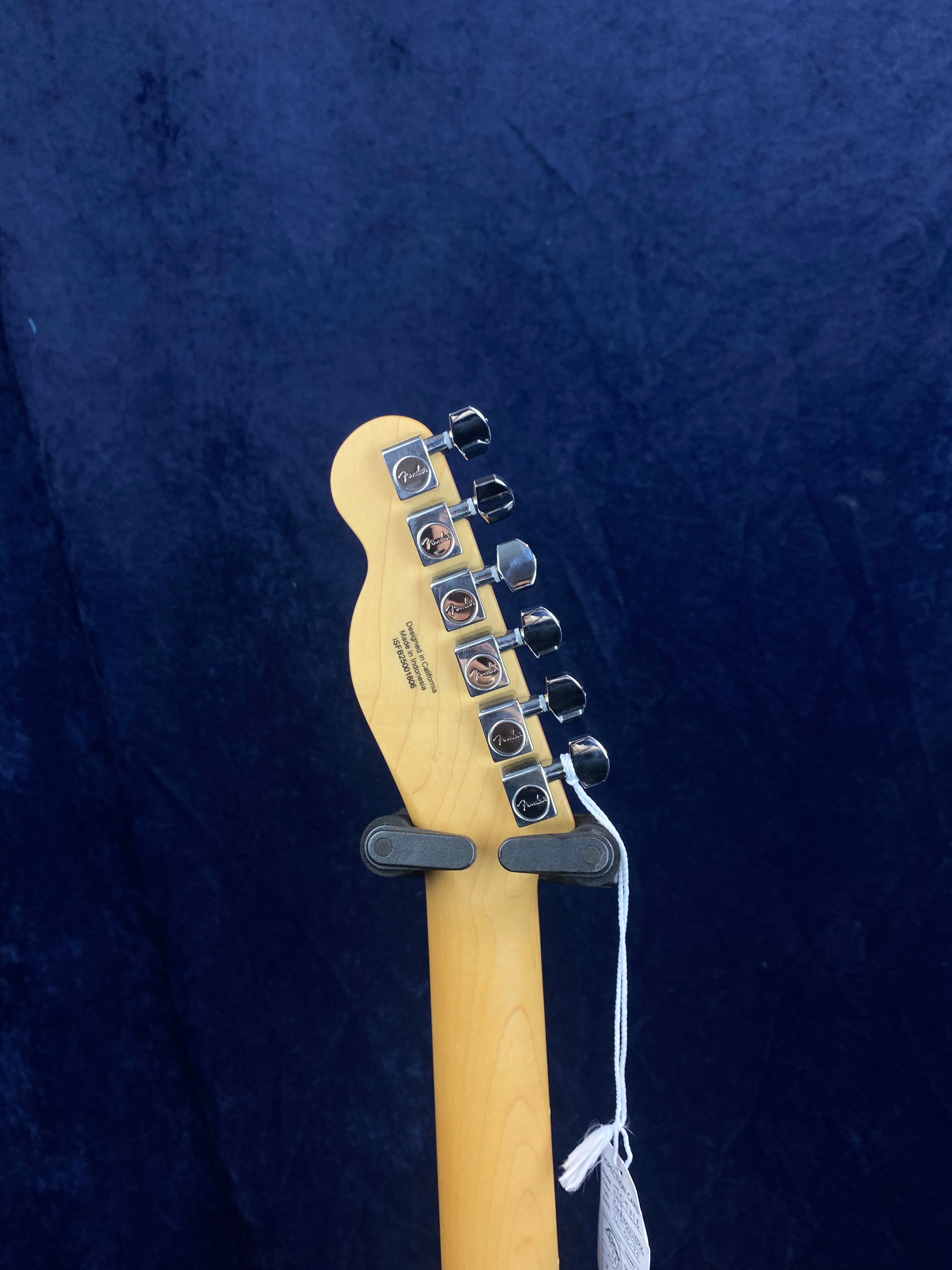 Six tuning pegs and the Fender logo appear on the headstock of a light wood electric guitar, shown from the back against a dark blue background.