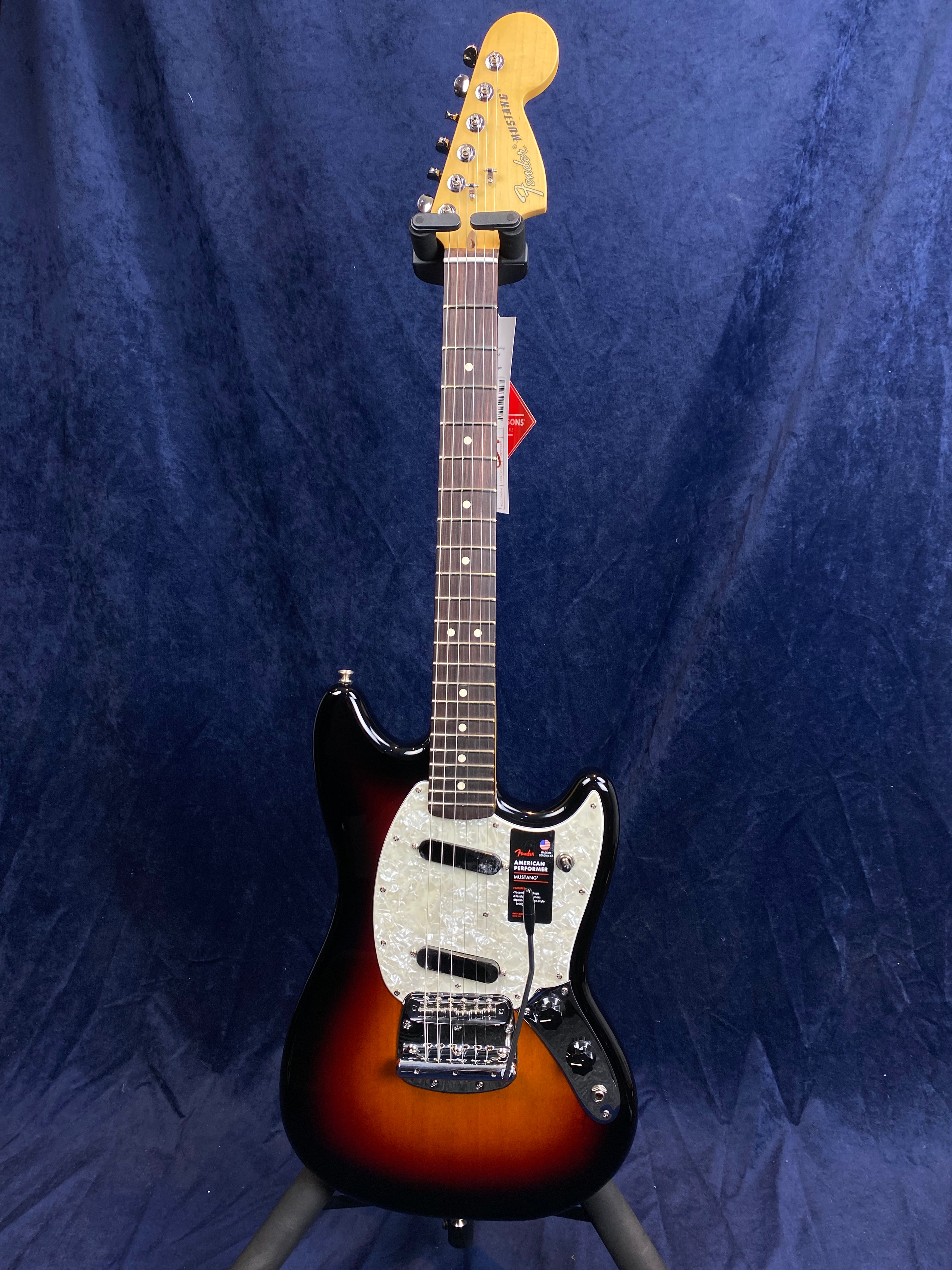 Sunburst Fender Mustang electric guitar with a white pearloid pickguard, dark rosewood fretboard, and chrome control knobs, standing on a tripod.