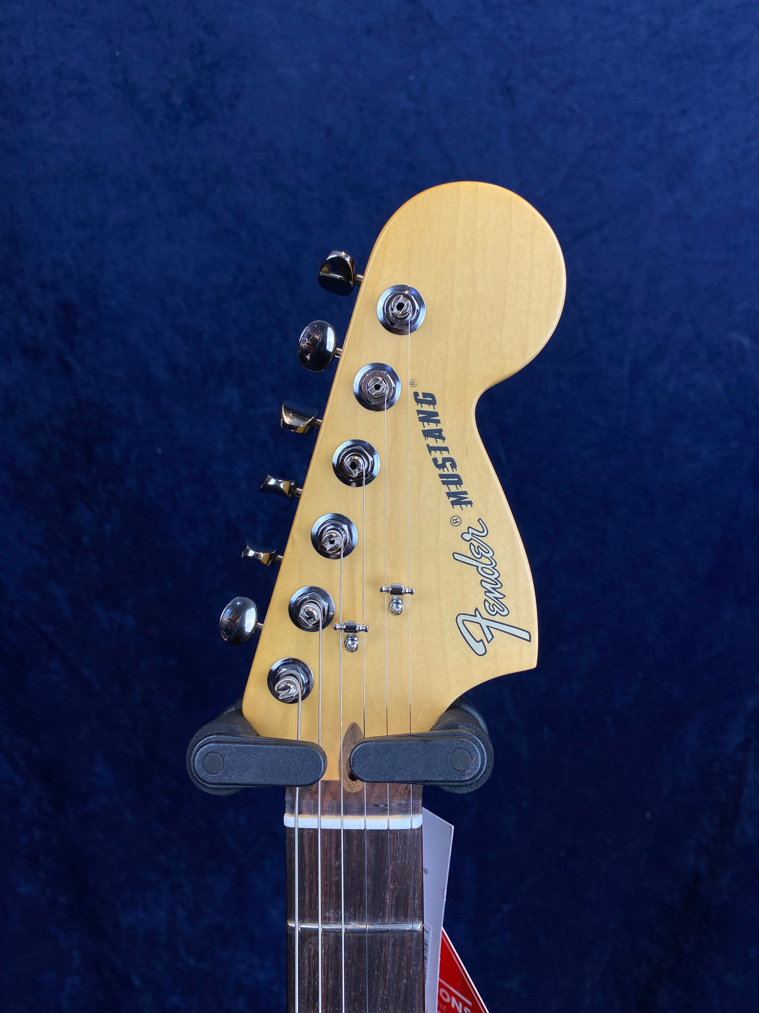 Natural wood Fender electric guitar headstock with six chrome tuning pegs, Fender logo, and dark fingerboard, positioned against a dark blue background.