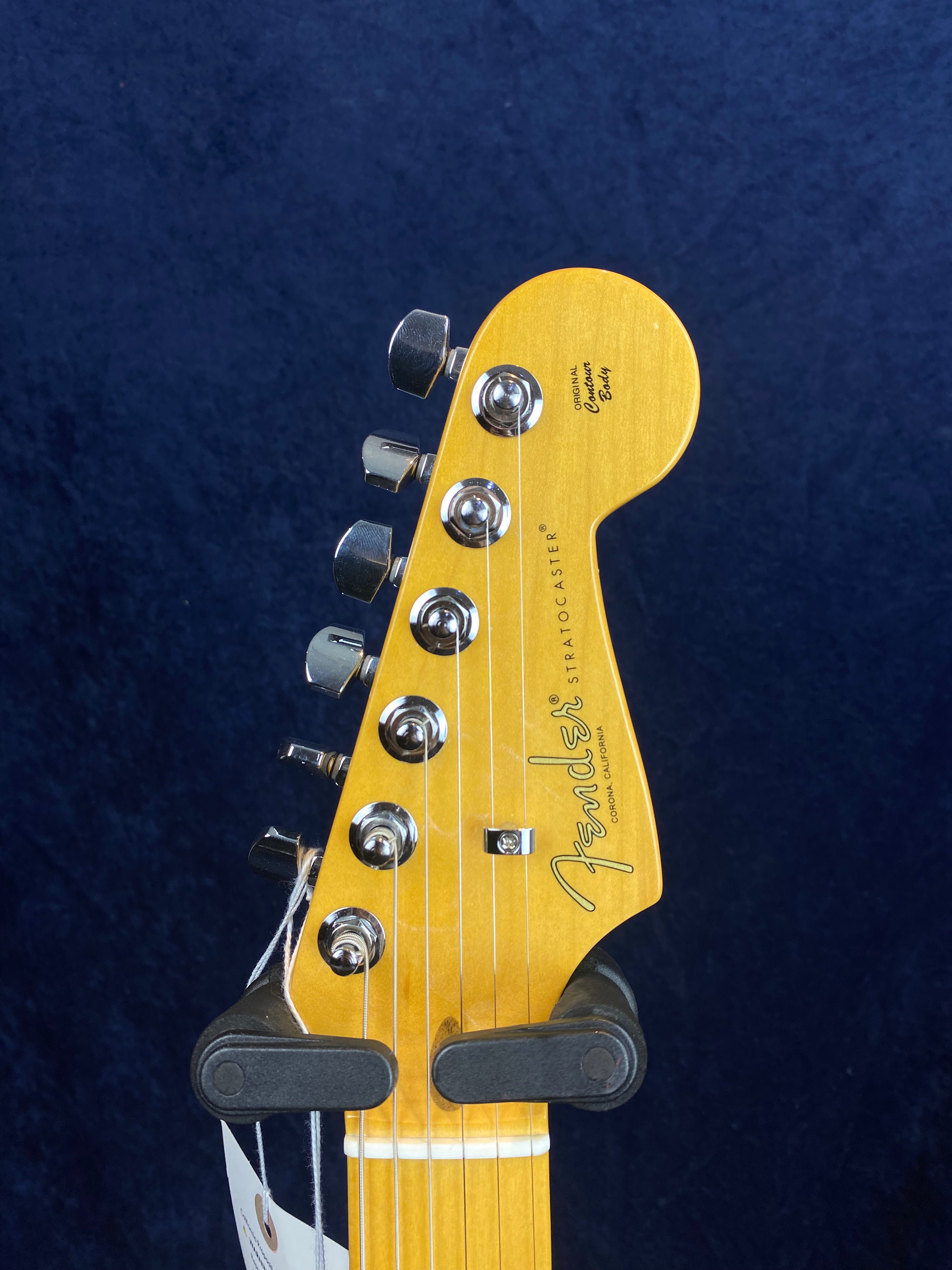 Light wood Fender Stratocaster electric guitar headstock with six chrome tuning pegs, string tree, and black Fender logo on blue background.