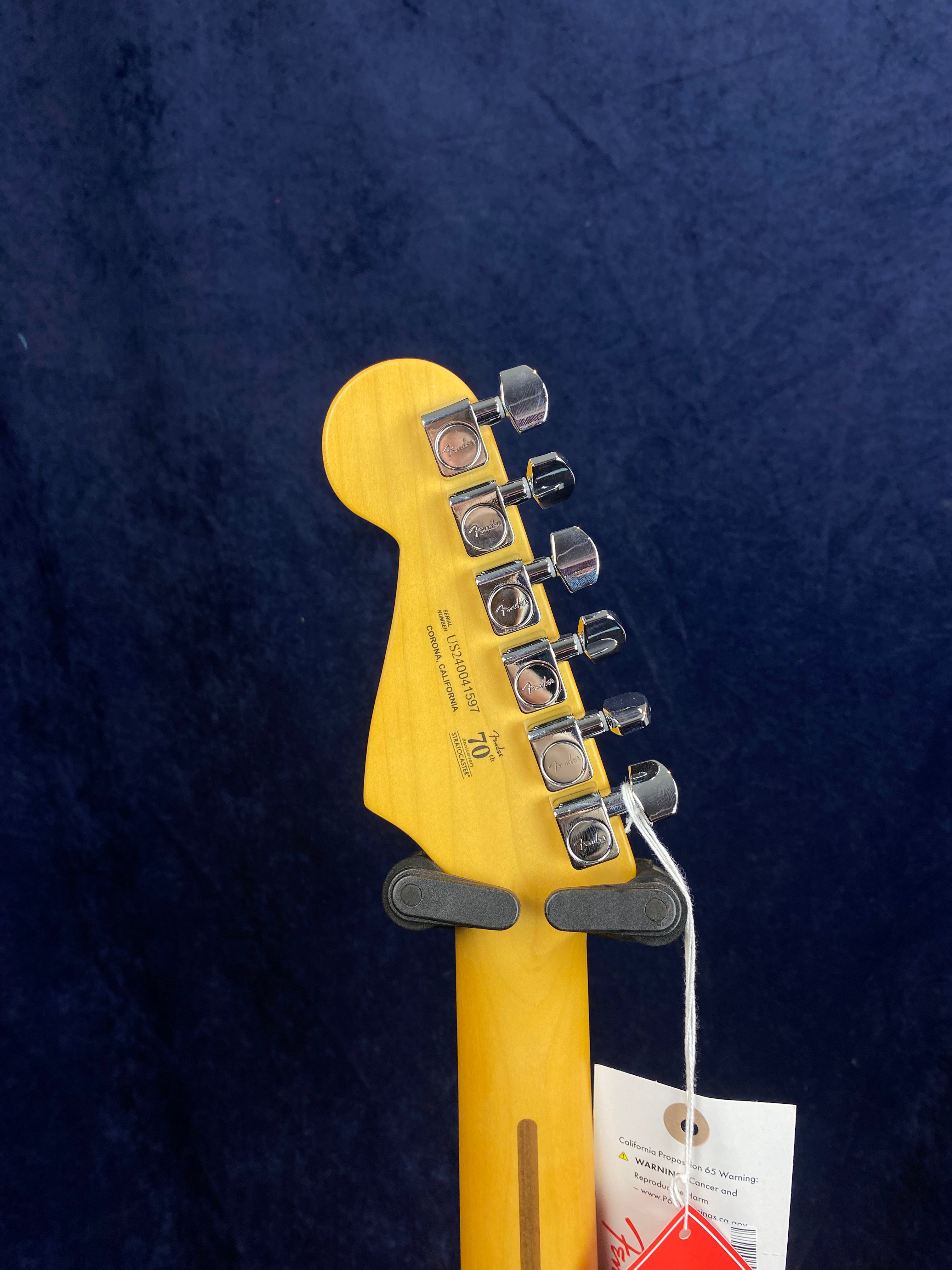 Headstock and tuners of a Fender electric guitar, with tags hanging from one tuner, against a dark blue background.