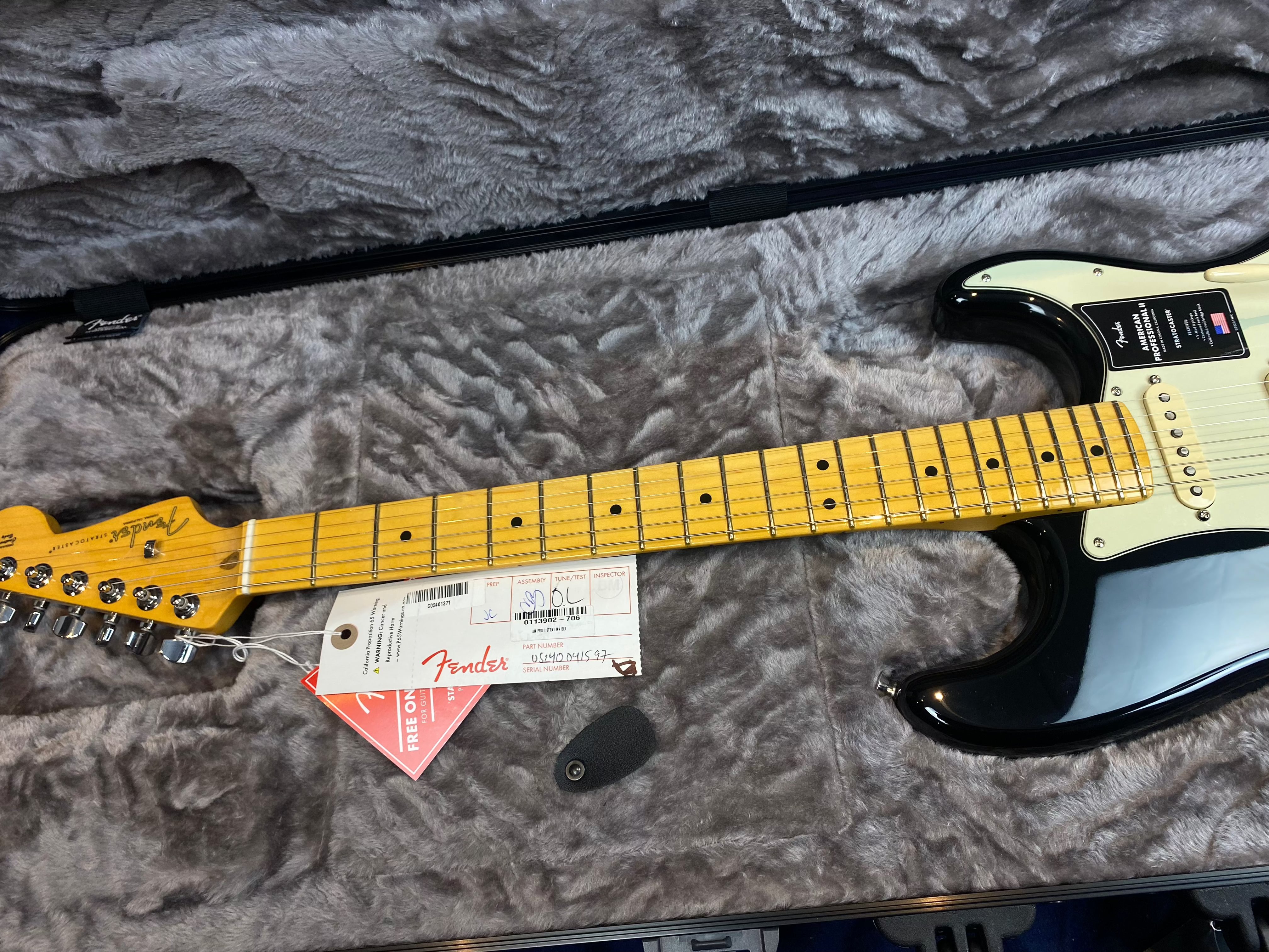 Black Fender electric guitar with a maple neck and fretboard, lying in a gray plush case, tags attached near the headstock, and a pick beside it.