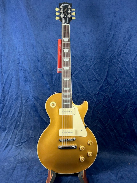 Gold-colored electric guitar with white pickups, pickguard, and control knobs, standing upright against a dark blue textured background.