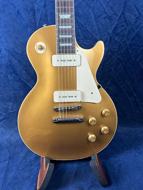 Gold electric guitar with white pickguard, two pickups, four gold control knobs, and a glossy finish, displayed upright on a stand.