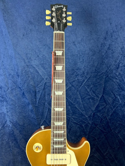 Gibson electric guitar with a gold top, cream pickguard, trapezoid inlays on the fretboard, and yellow tuning pegs, shown against a blue background.