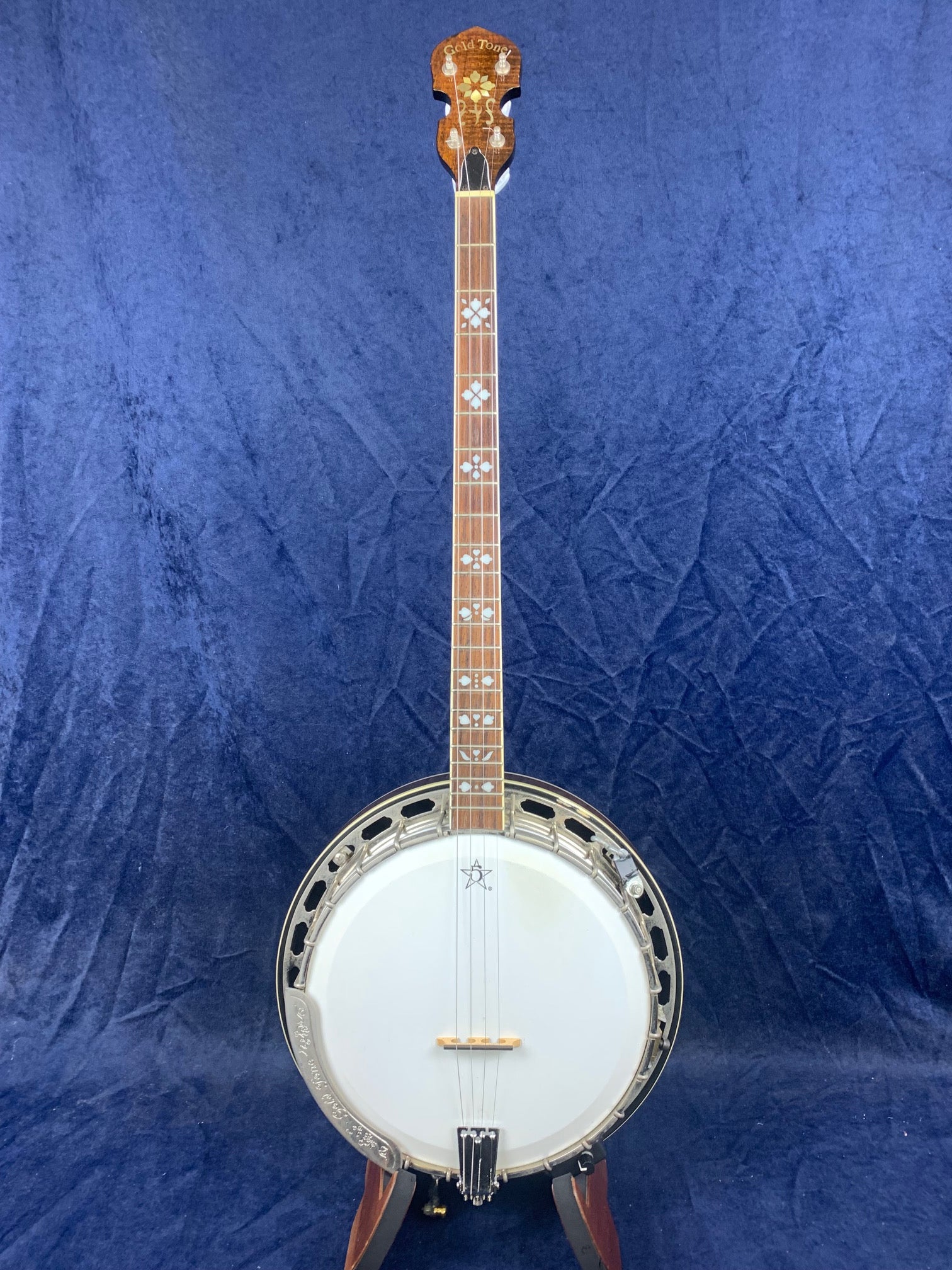 Vintage five-string banjo with a wood neck, decorative inlays on the fretboard, metal rim, and white drumhead, standing upright against a blue cloth backdrop.